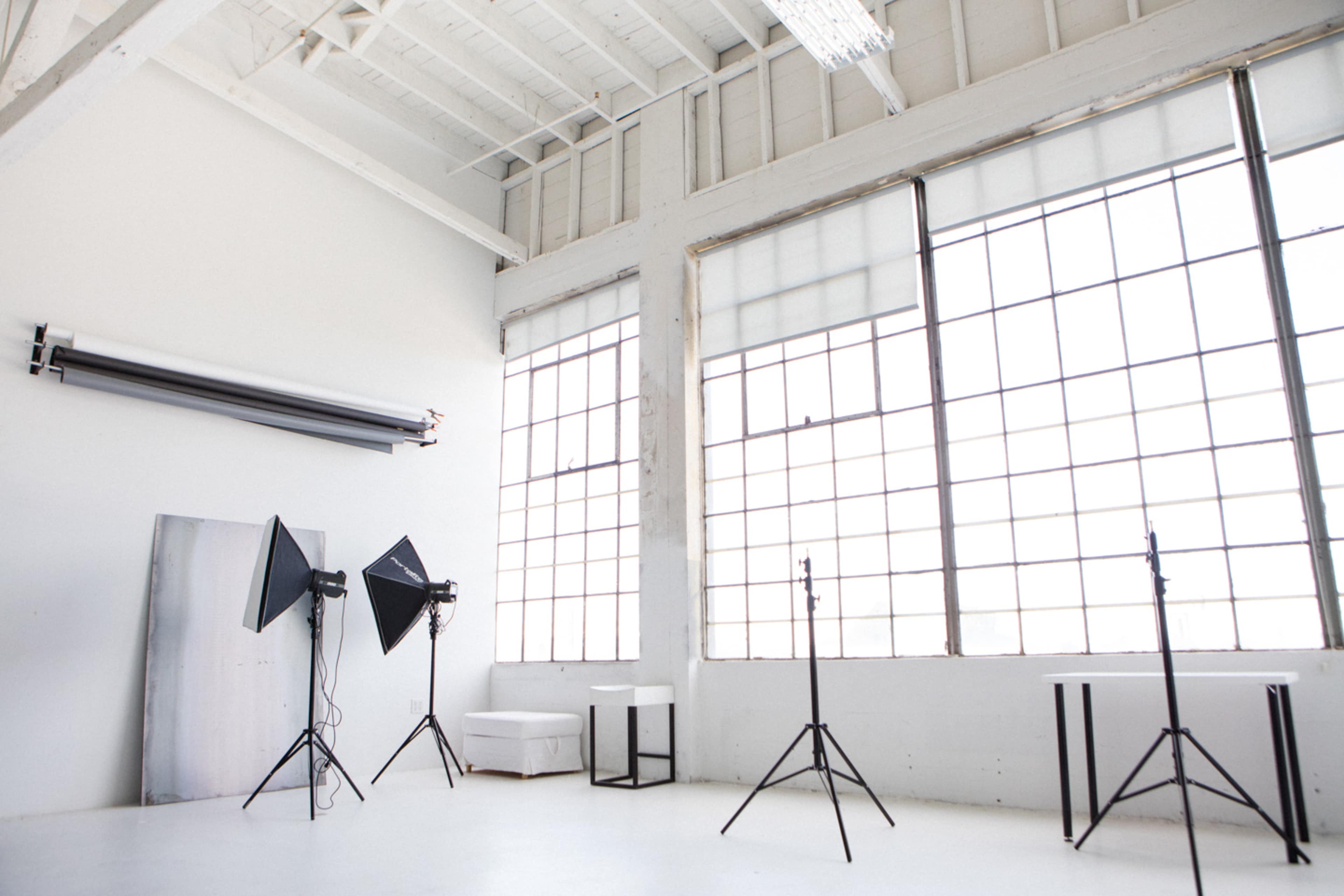A spacious photo studio featuring large windows, backdrop rolls, and lighting equipment set up on stands.