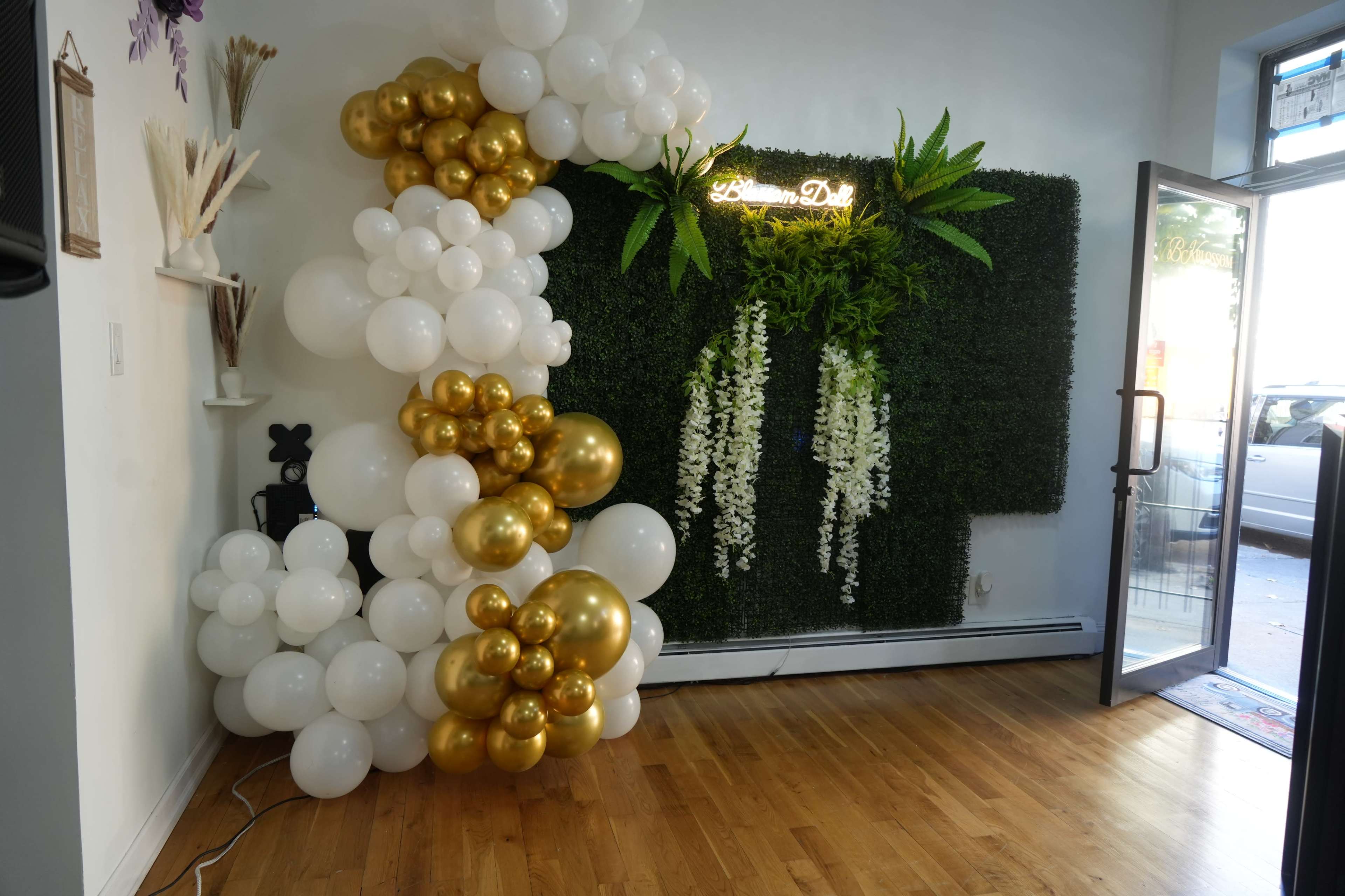 A corner of a decorated room featuring a large arrangement of white and gold balloons next to a green wall with hanging plants and a neon sign.