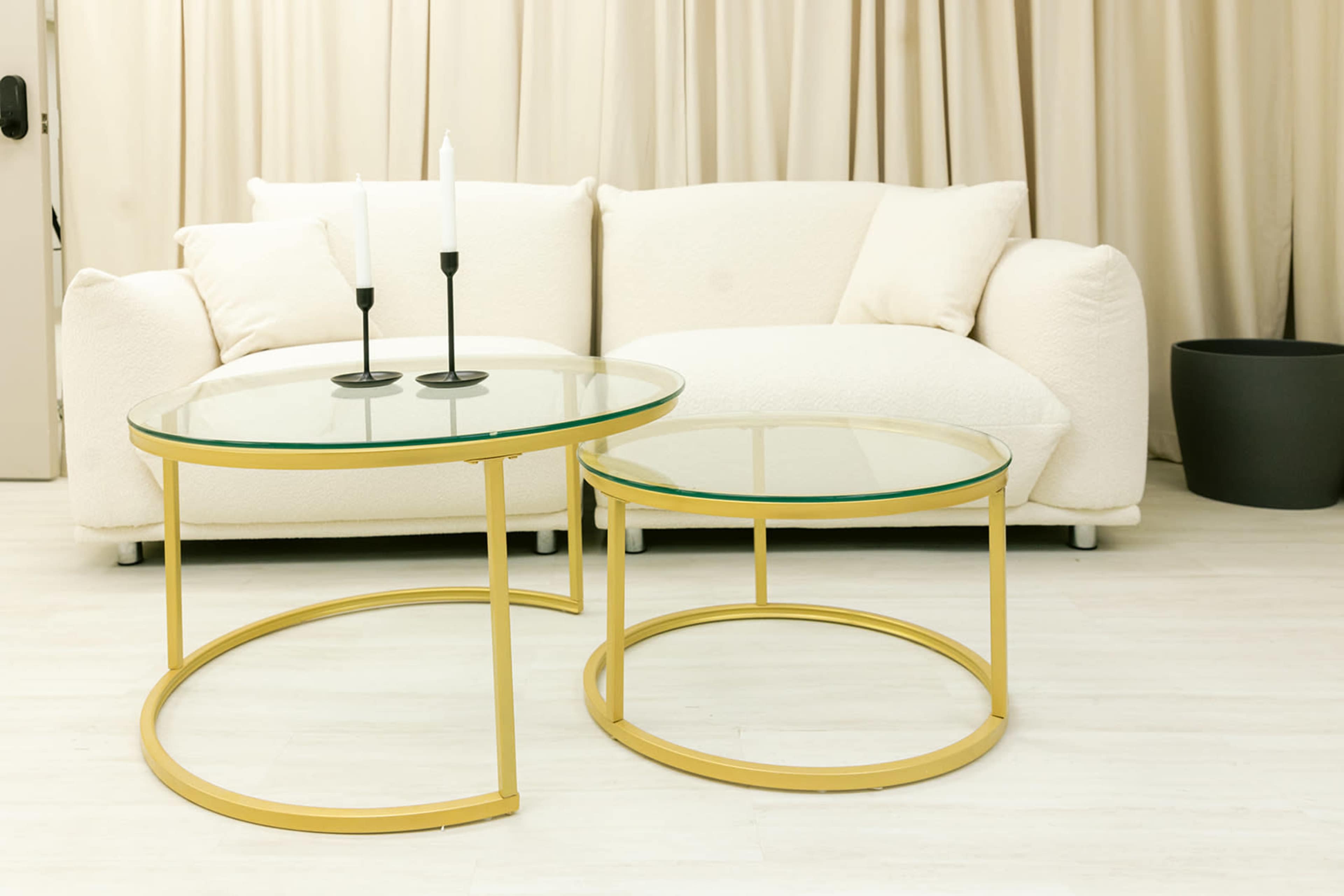 A white couch is positioned against a backdrop of beige curtains, accompanied by two circular glass-top coffee tables with gold frames and a black planter nearby.