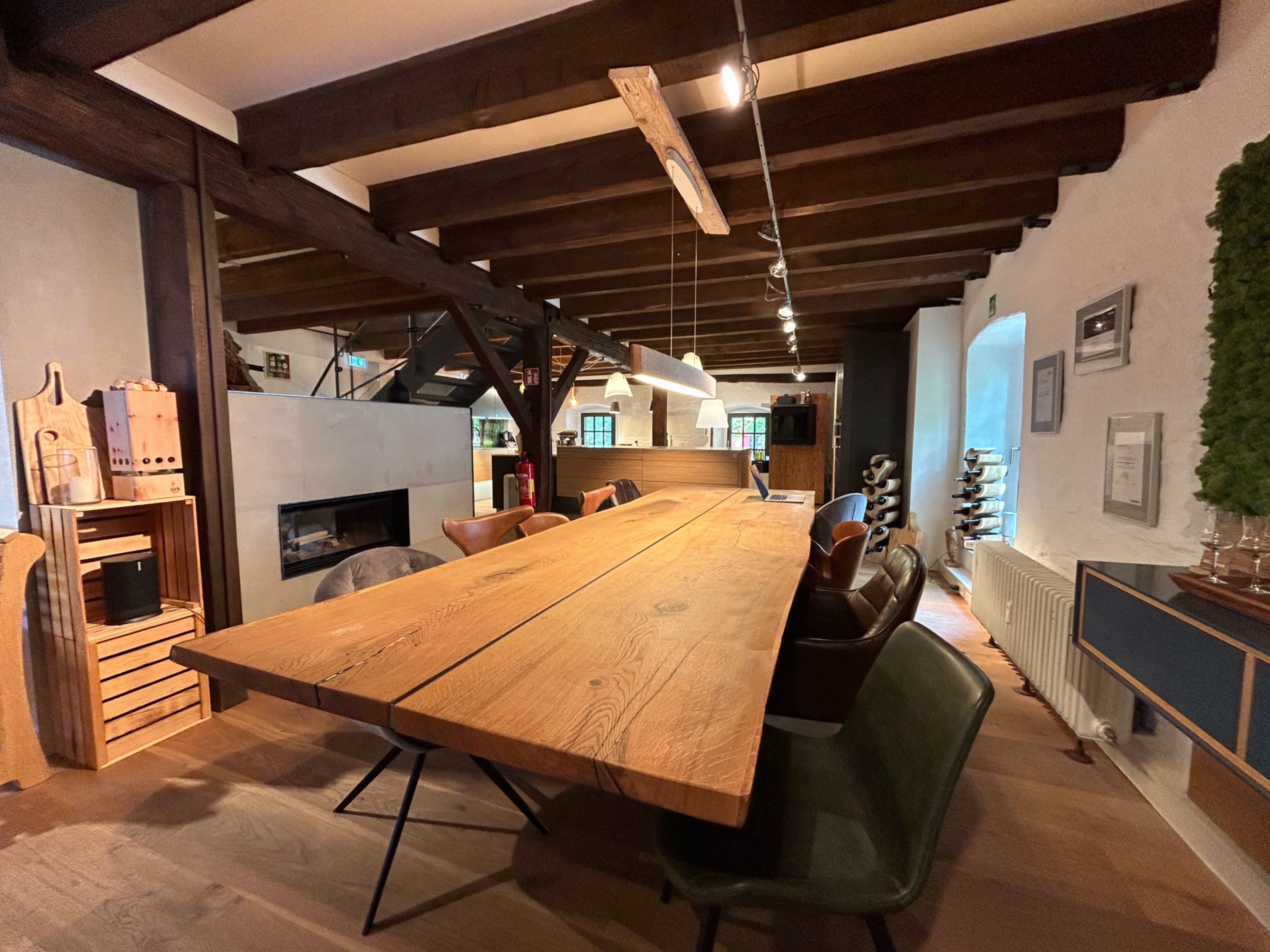 The image shows a spacious dining area with a large wooden table, wooden beams in the ceiling, and modern decor elements.