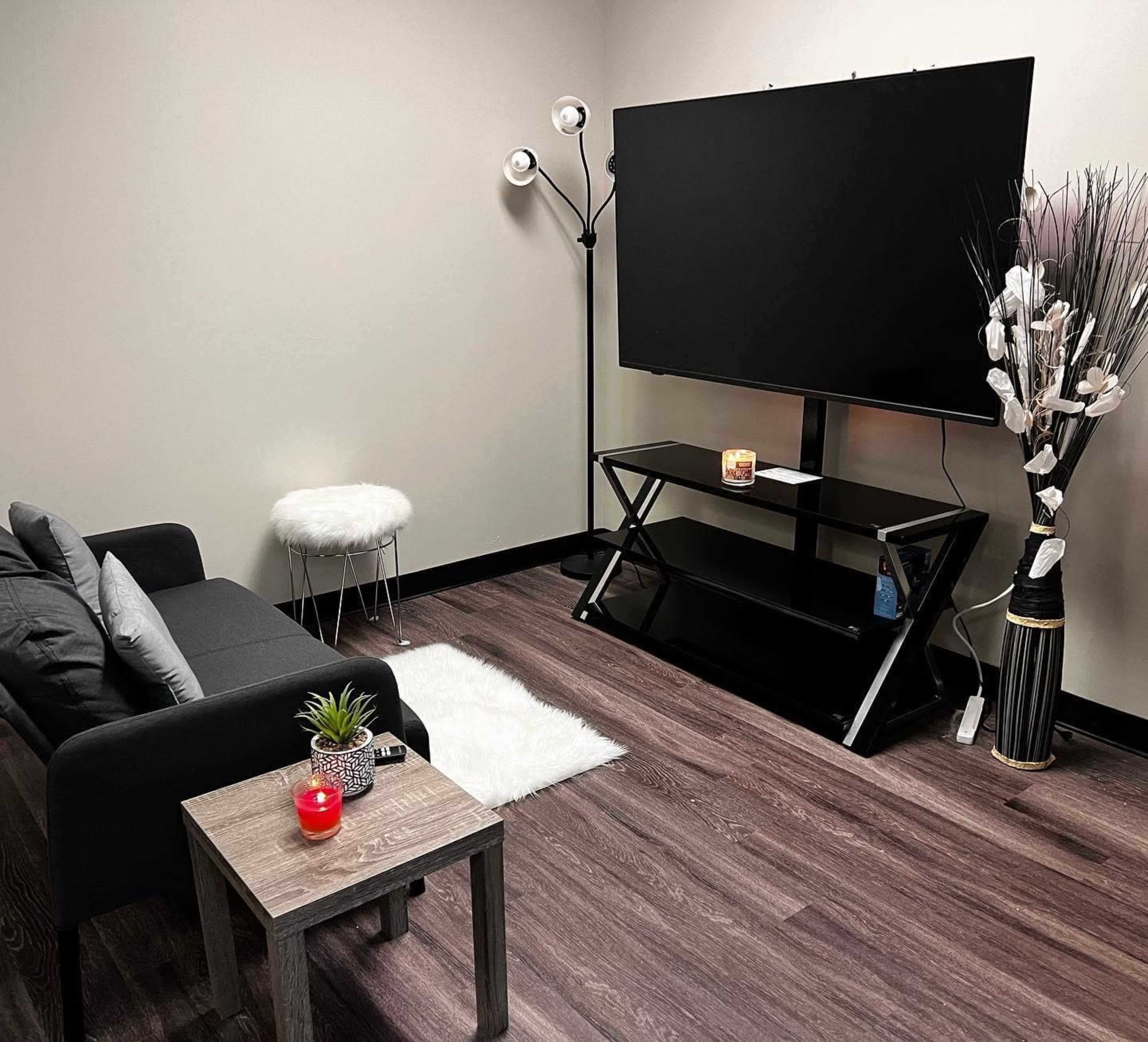 The image shows a small, modern room featuring a black couch, a coffee table with a plant and a candle, a television mounted on the wall, and a decorative floor lamp.