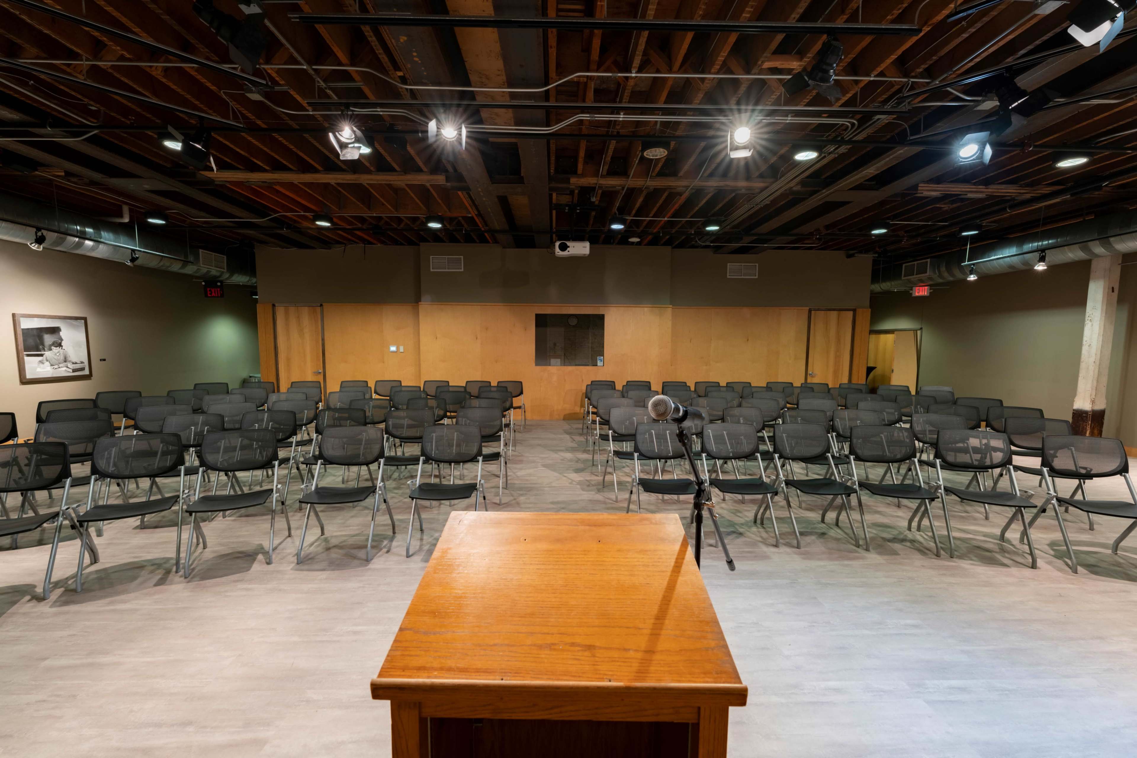 The image shows a semi-empty auditorium with rows of metal folding chairs facing a wooden podium under dim ceiling lights.