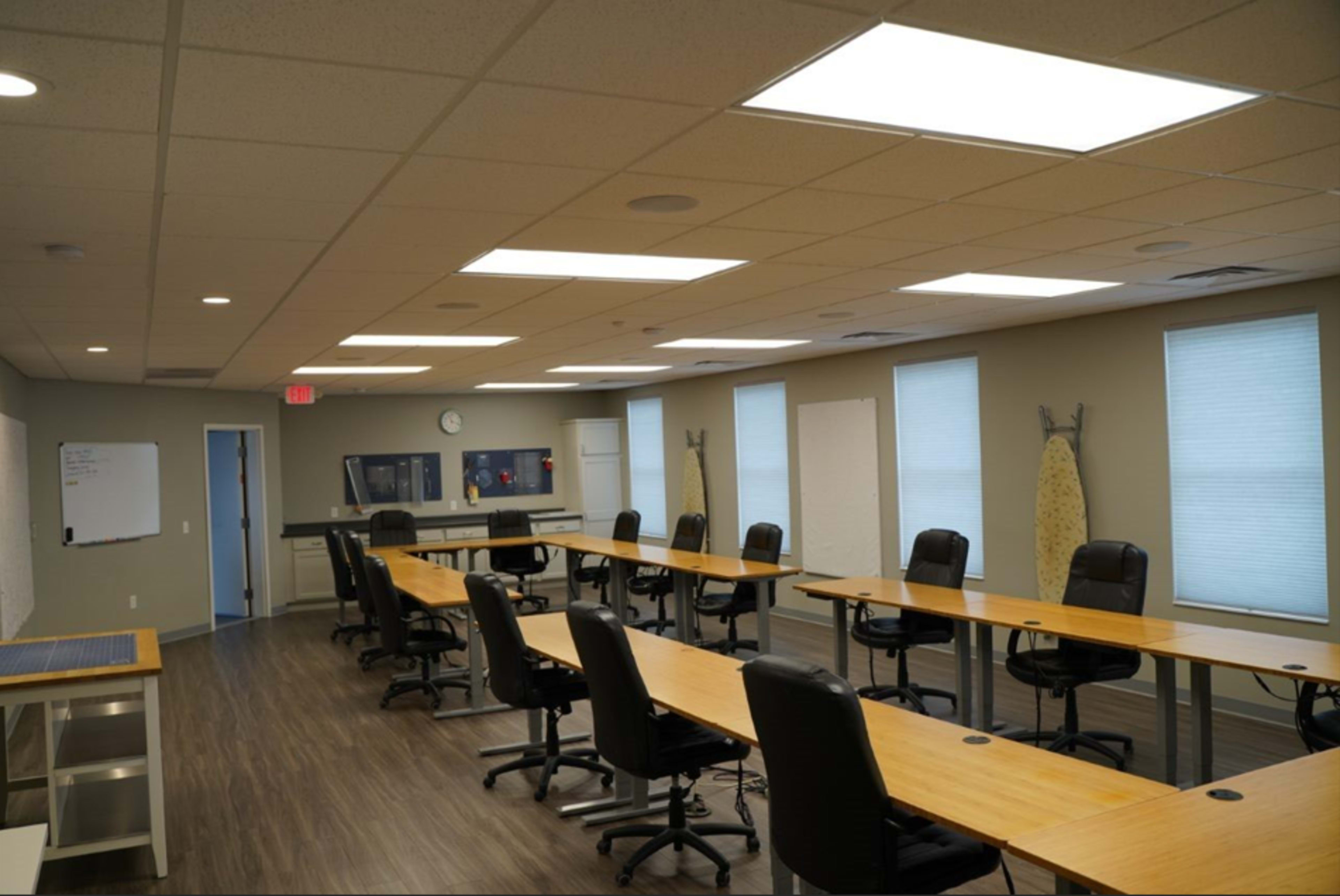 A well-lit classroom features several long tables and black rolling chairs arranged in a spacious layout, with whiteboards and windows along the walls.