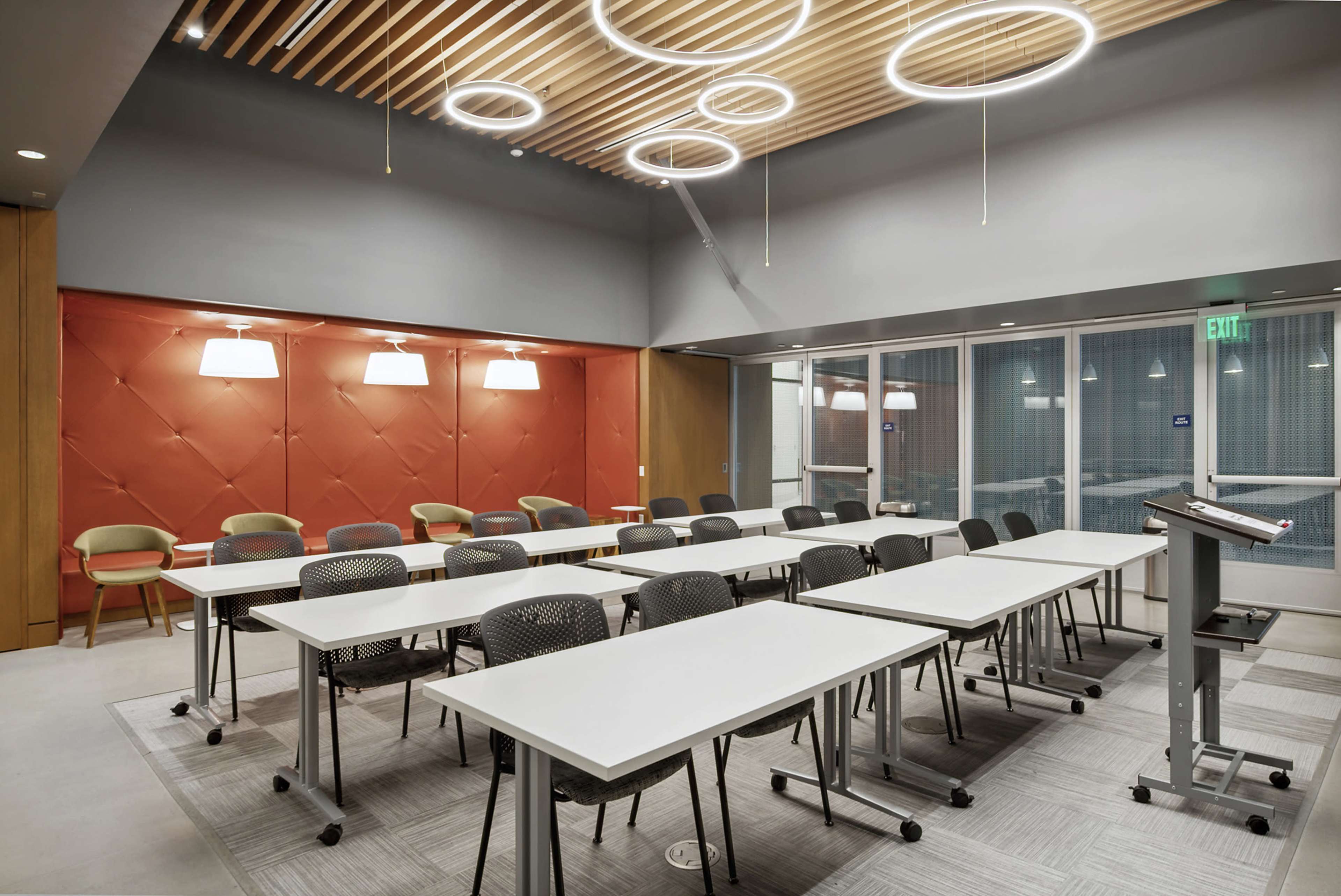 A modern classroom set up with multiple rows of white tables and black chairs, illuminated by circular light fixtures and featuring a red accent wall.
