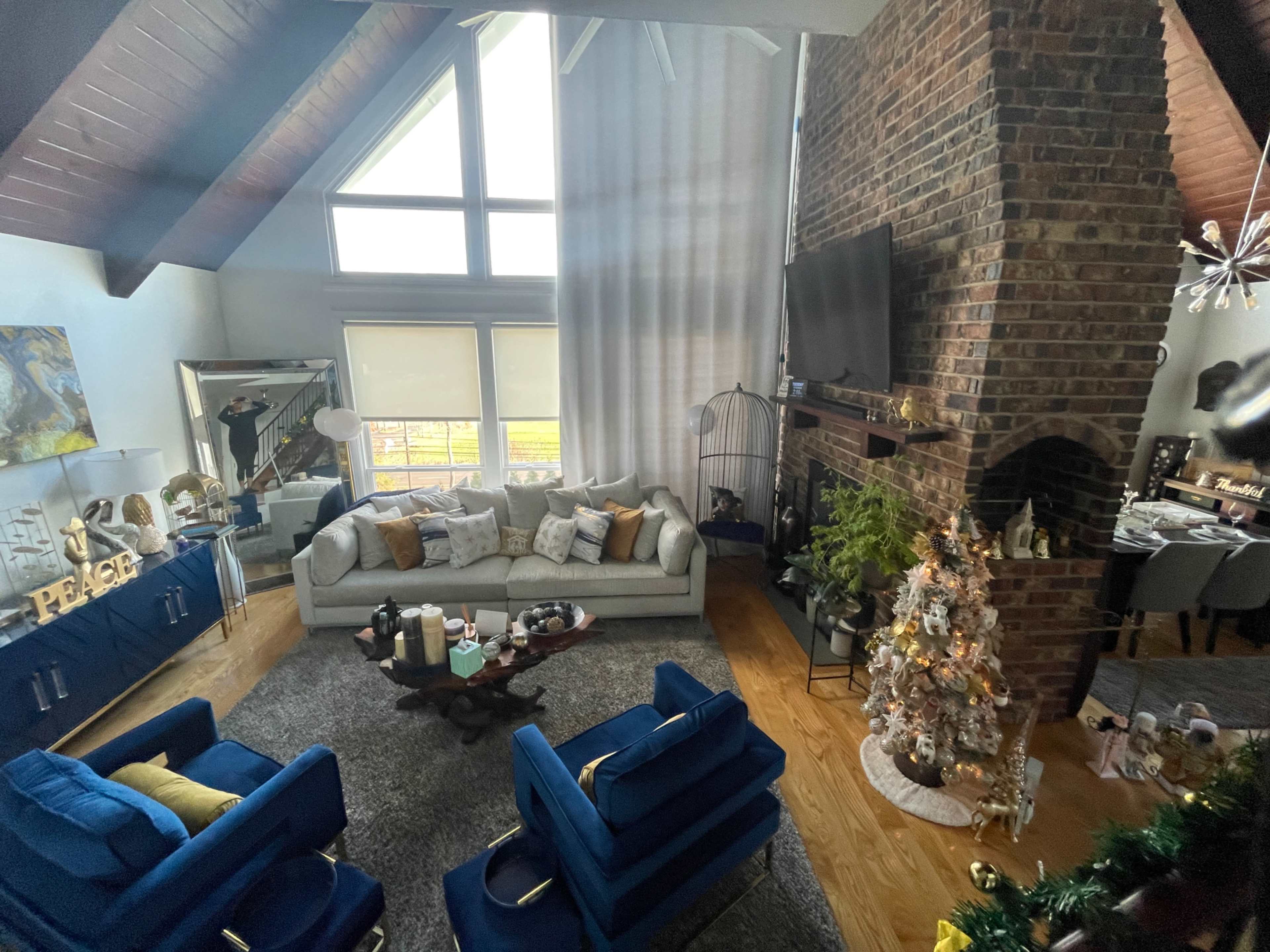 The image shows a spacious living room featuring a large window, a light-colored sofa, blue accent chairs, a Christmas tree, and a brick fireplace.