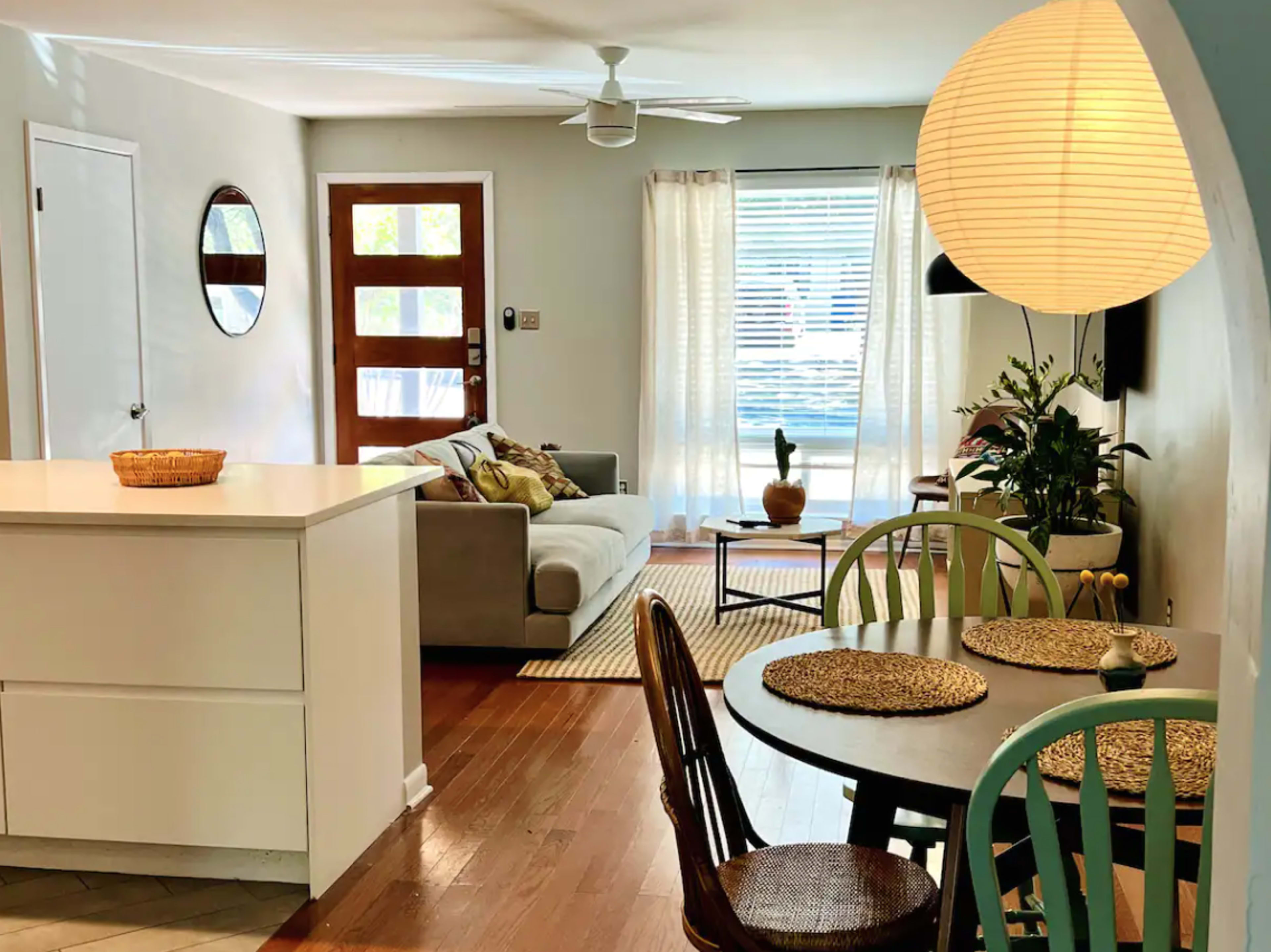 The image shows a cozy living space with a sofa, a round dining table, and a large window that lets in natural light.