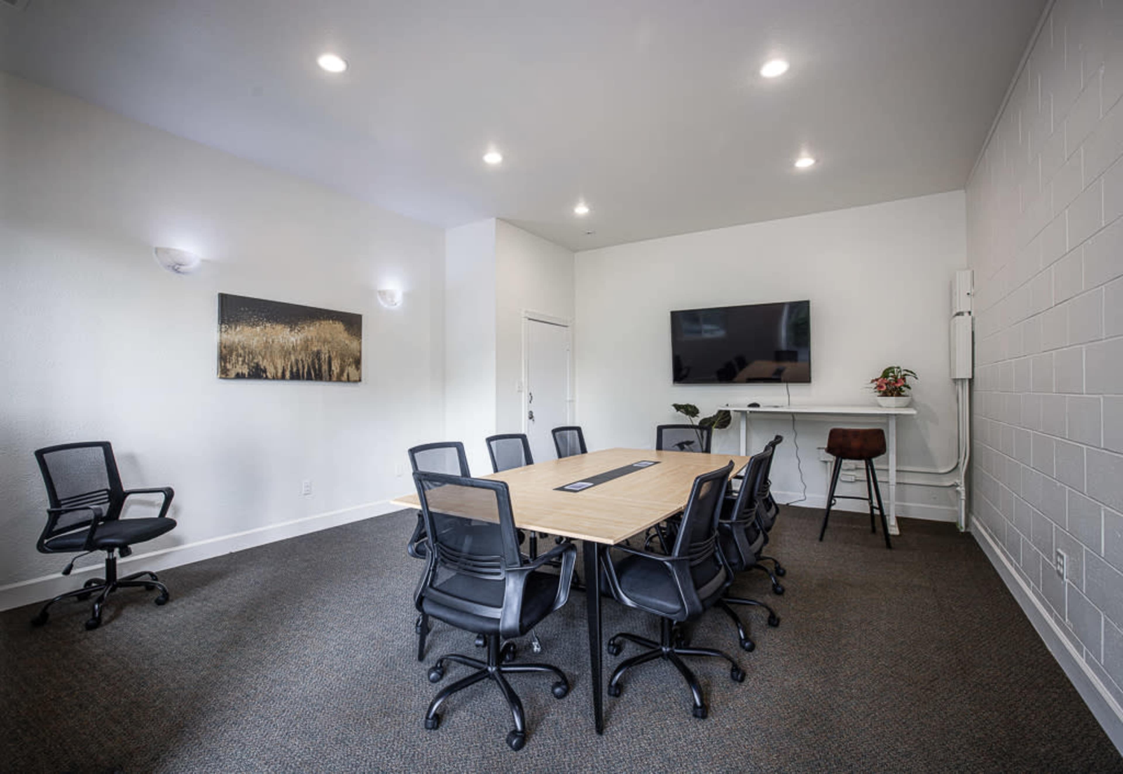 A modern conference room with a long wooden table surrounded by black chairs, a television mounted on the wall, and a small plant on a shelf.