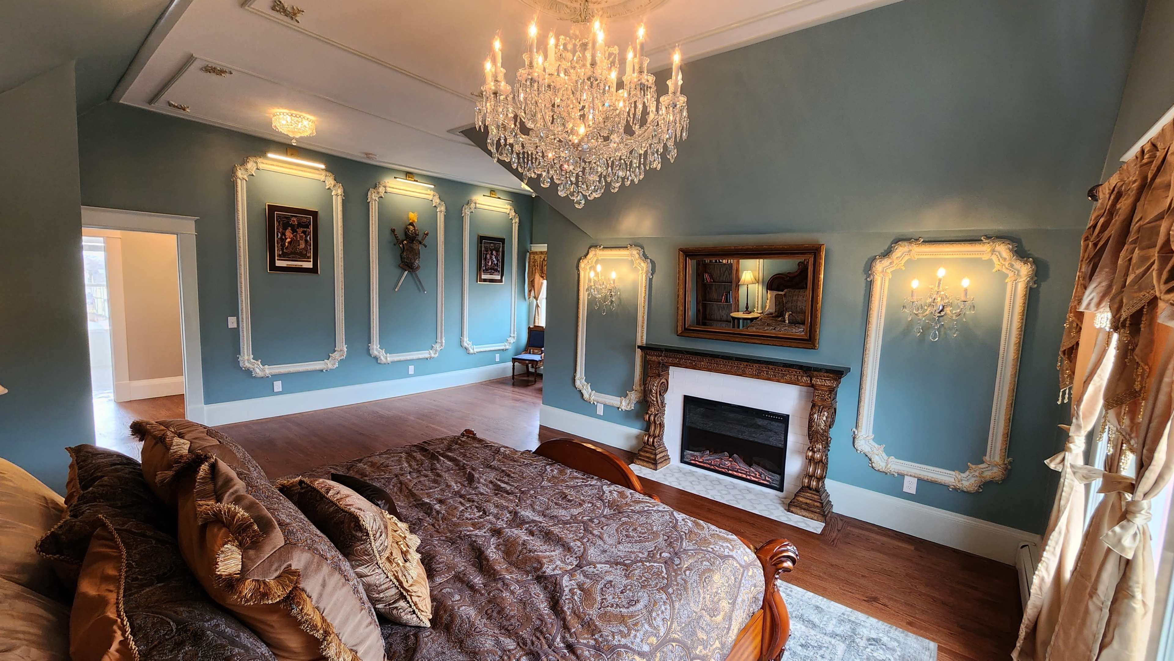 A decorated bedroom featuring a chandelier, ornate wall frames, a fireplace, and a large bed with decorative bedding.