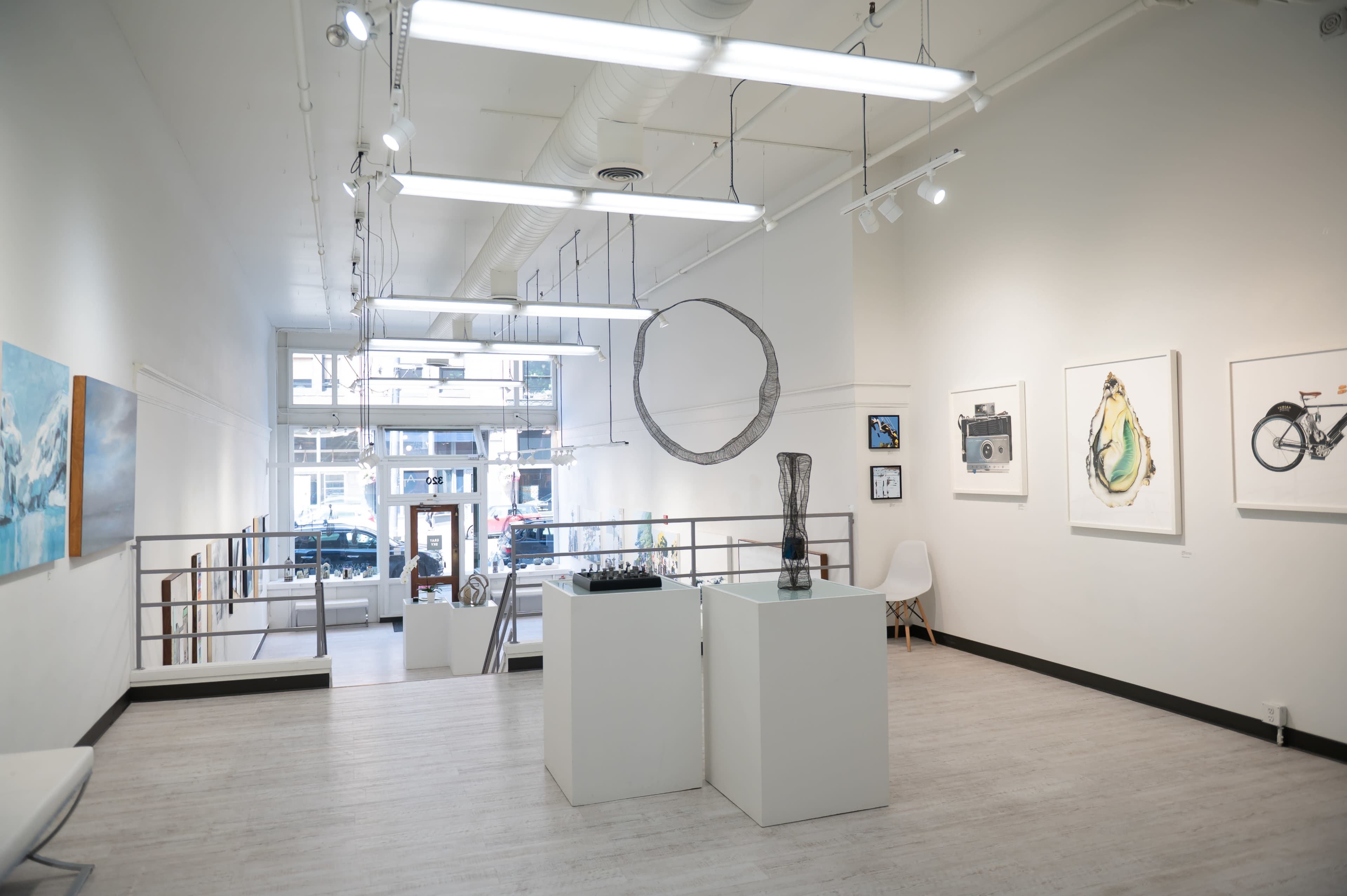 An art gallery with white walls, featuring various artworks displayed on the walls and sculptures on pedestals, with large windows allowing natural light to enter.