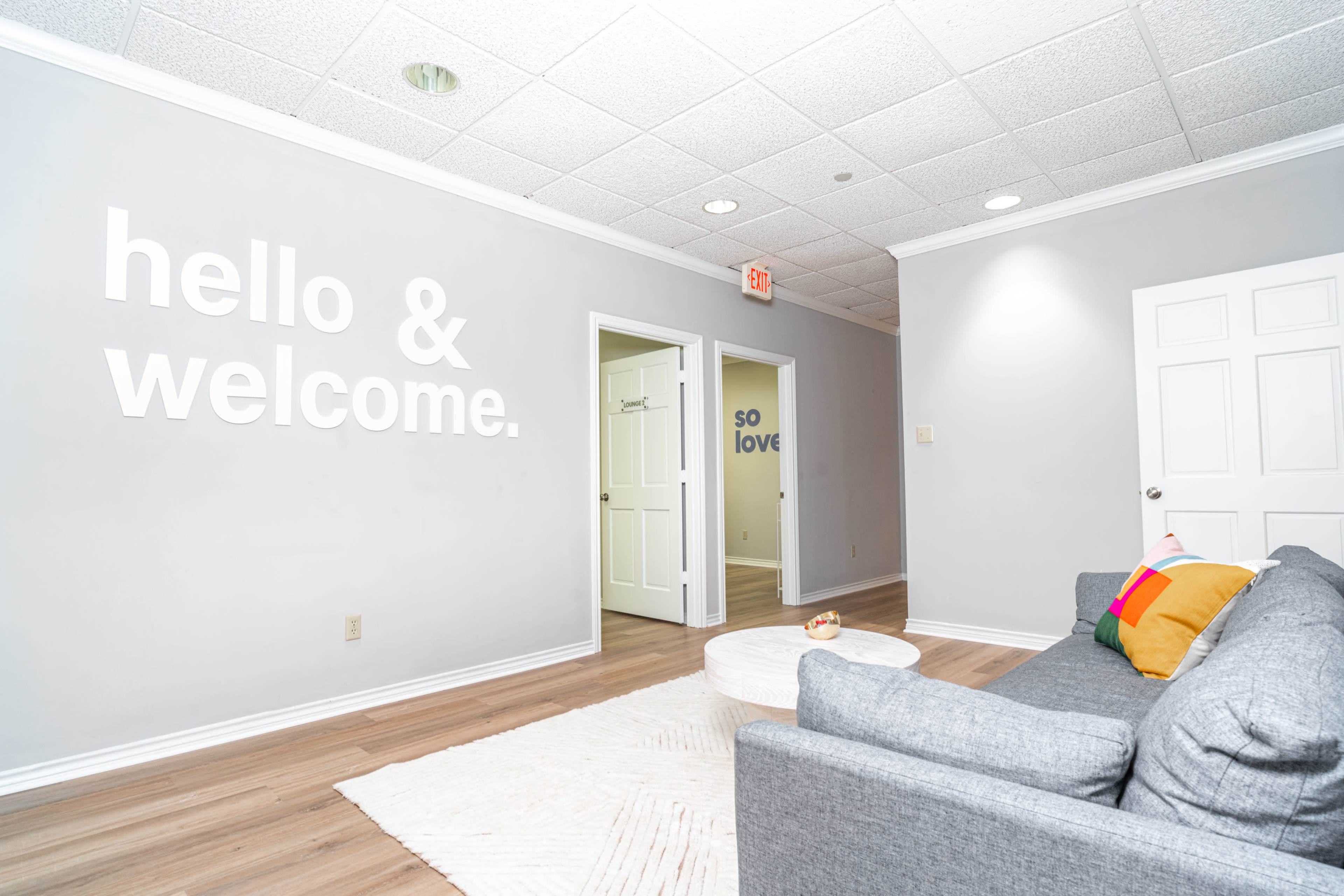 A modern, brightly lit living space with a gray sofa, a round coffee table, and welcoming wall decor featuring the phrases "hello & welcome" and "so love."