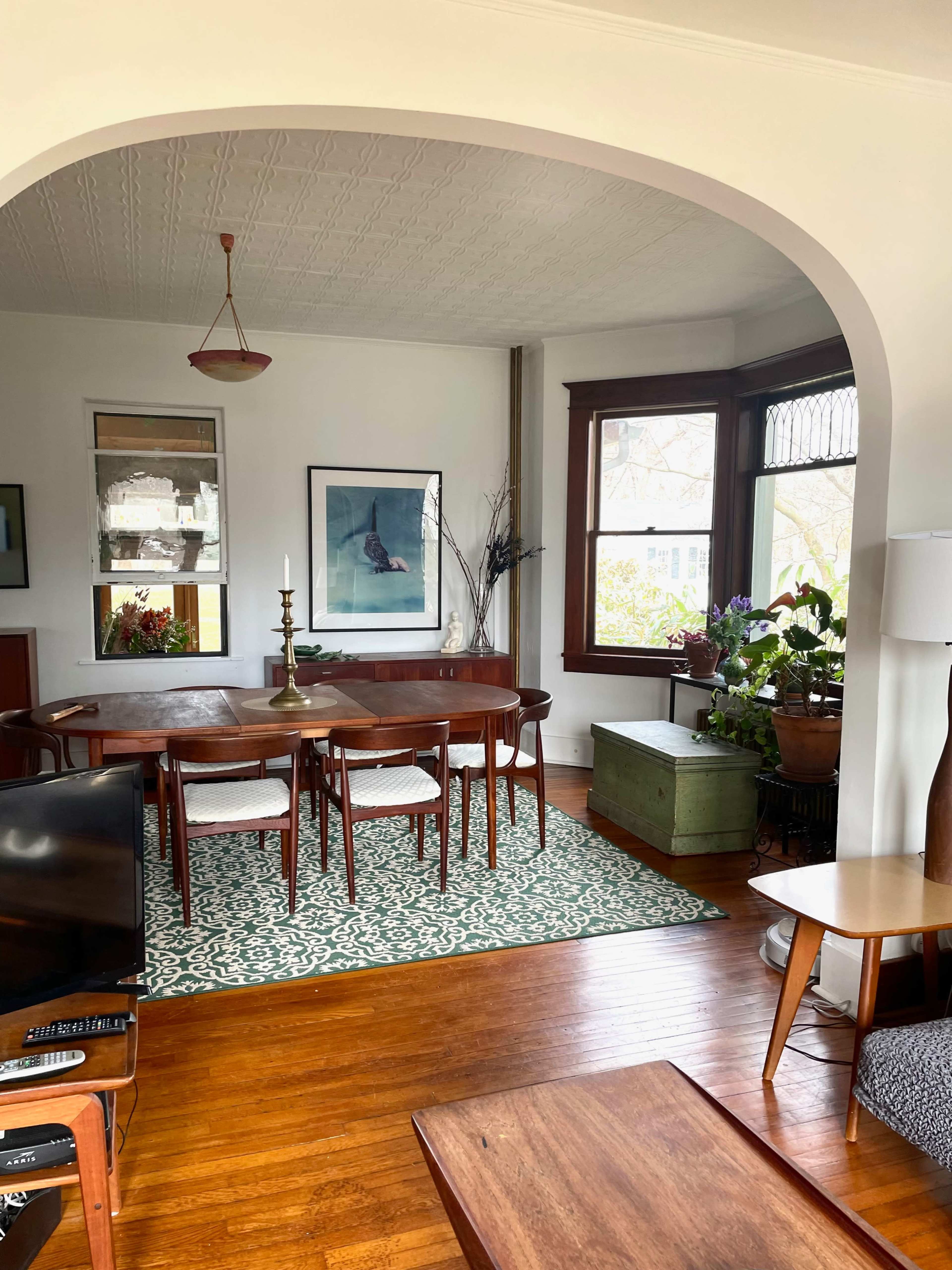 A dining area features a wooden table surrounded by chairs, framed artwork on the walls, and sunlight streaming through large windows.