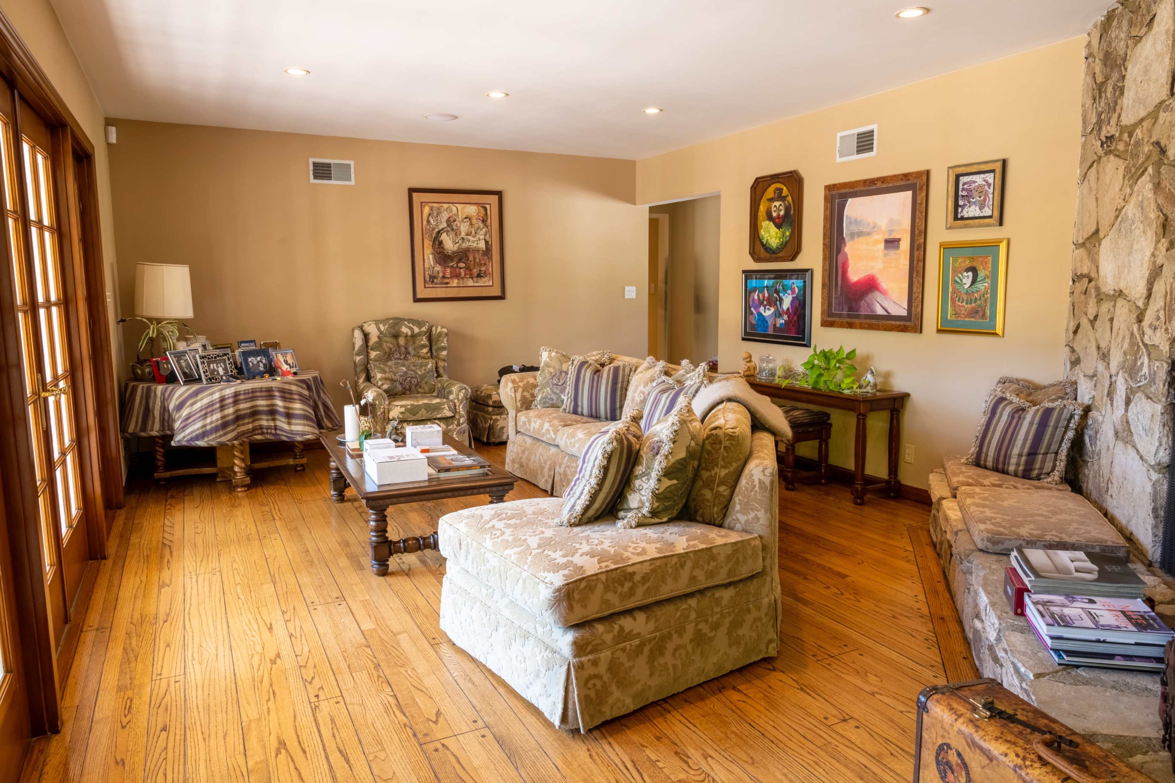 A living room with wooden flooring, a stone fireplace, and various framed pictures on the walls, featuring comfortable seating arranged around a coffee table.