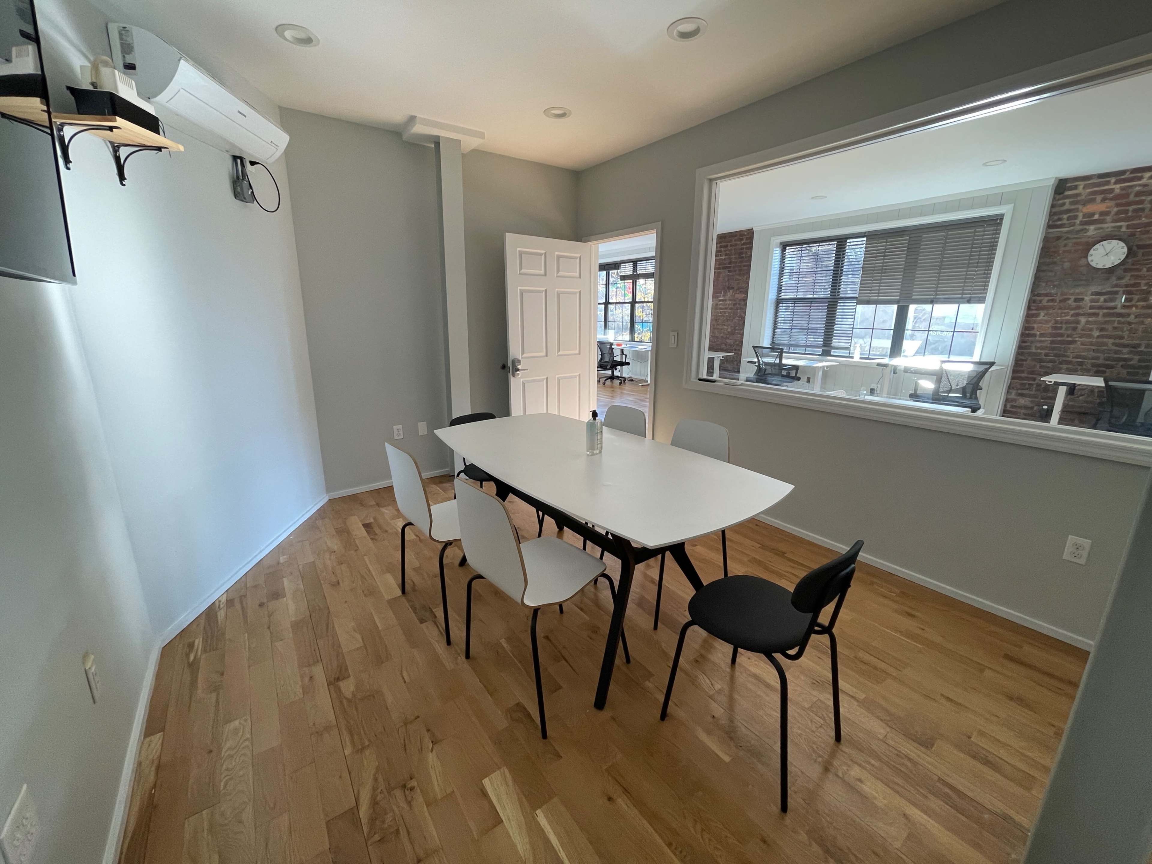 A modern meeting room with a white table surrounded by six chairs and large windows overlooking an office area.