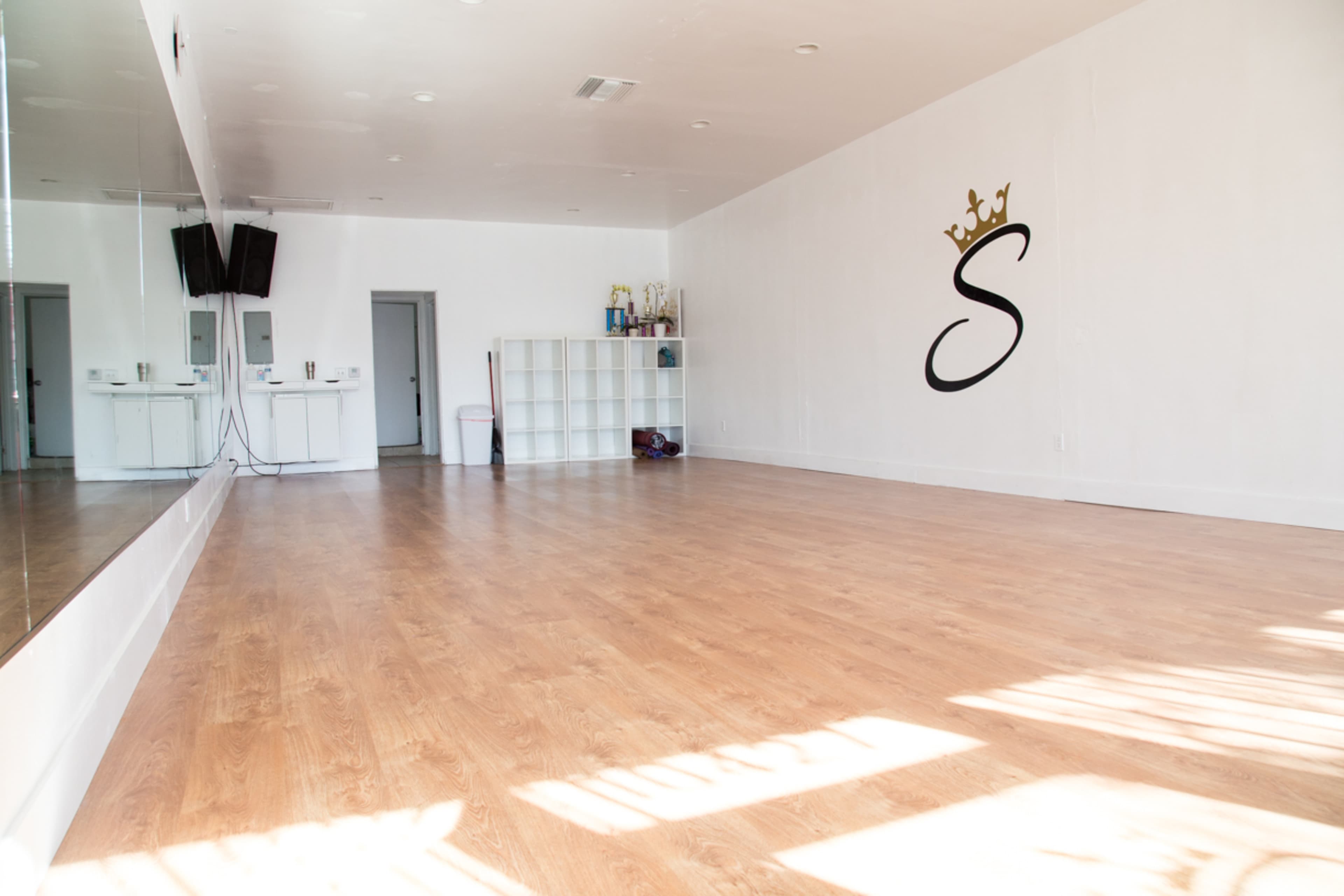 The image depicts an empty dance studio with polished wood flooring, a large mirror on one wall, and a logo featuring a crown and the letter "S" on the opposite wall.