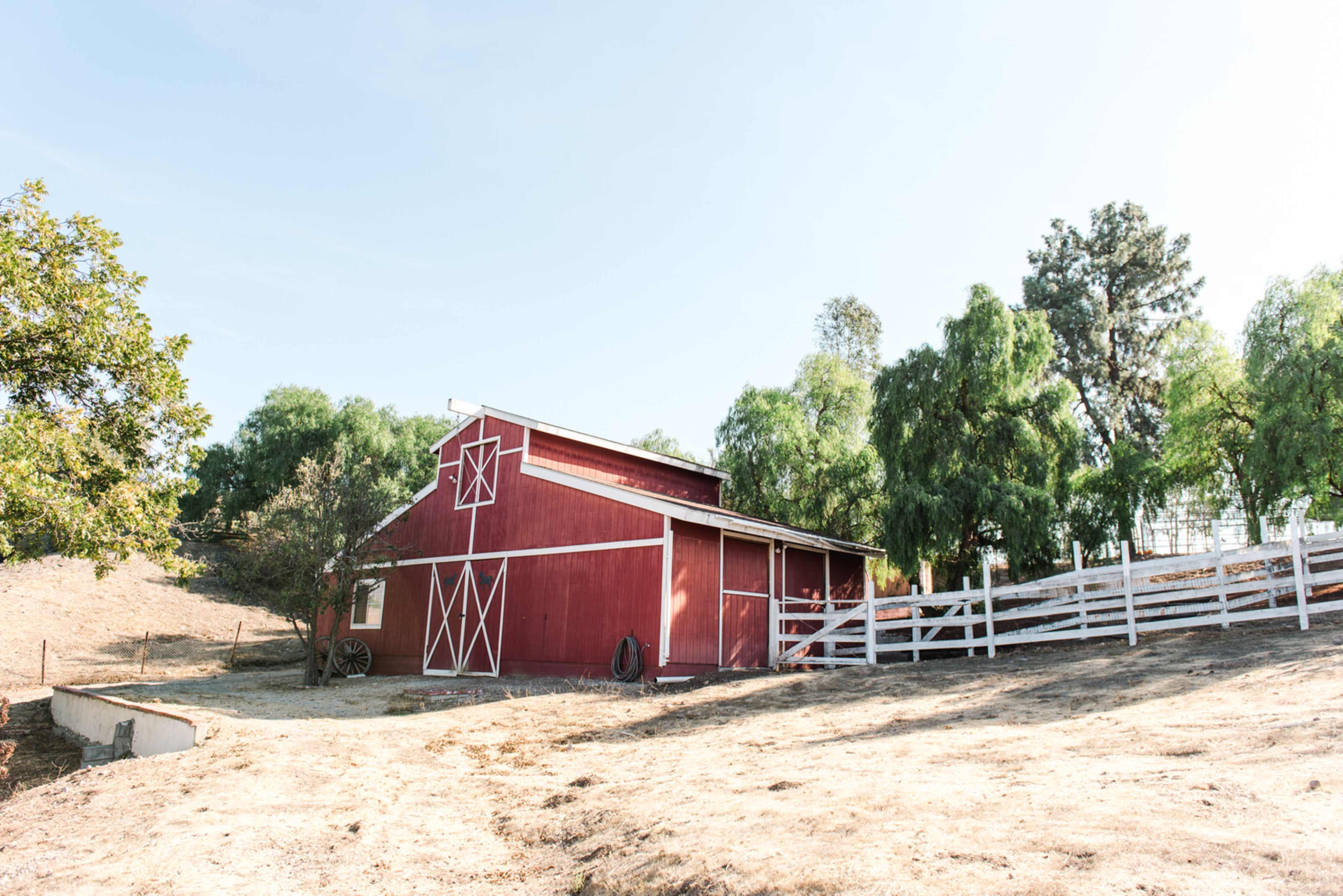 A red barn with white trim stands on a hillside surrounded by trees and a white fence.