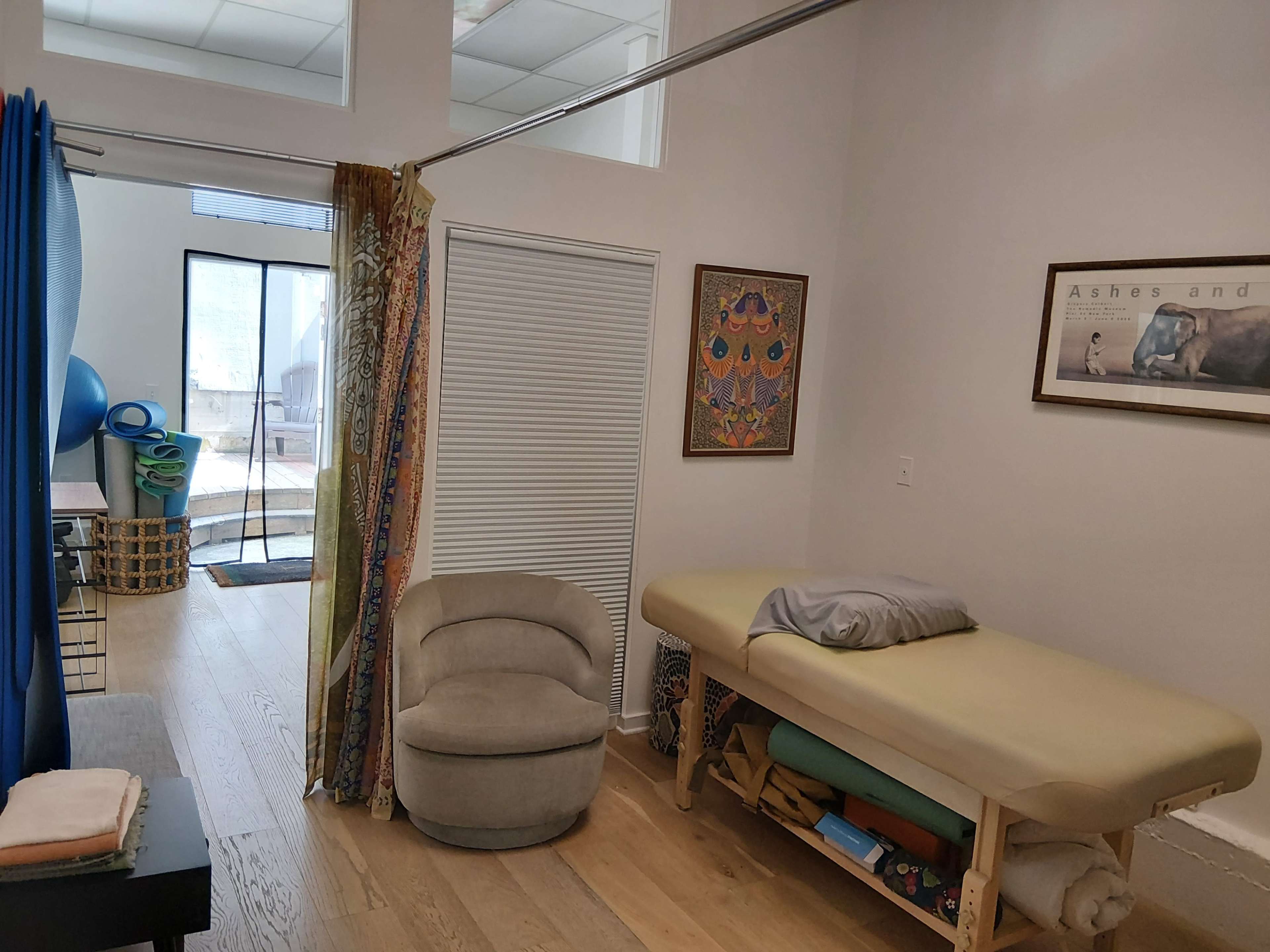 The image shows a therapy room with a massage table, a chair, and a partially open door leading to a light-filled outdoor area.