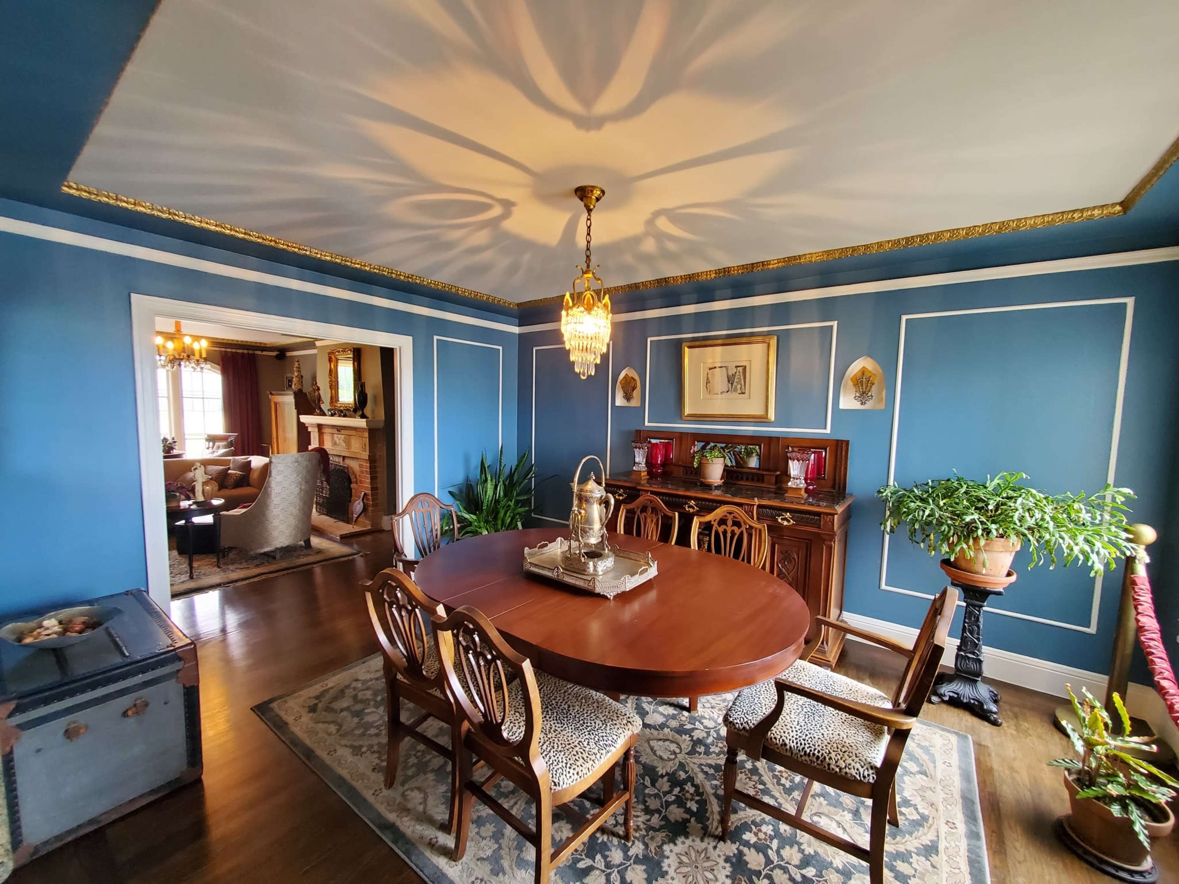 The image shows a dining room with a wooden table surrounded by chairs, a chandelier hanging from the ceiling, and blue walls adorned with decorative moldings and plants.