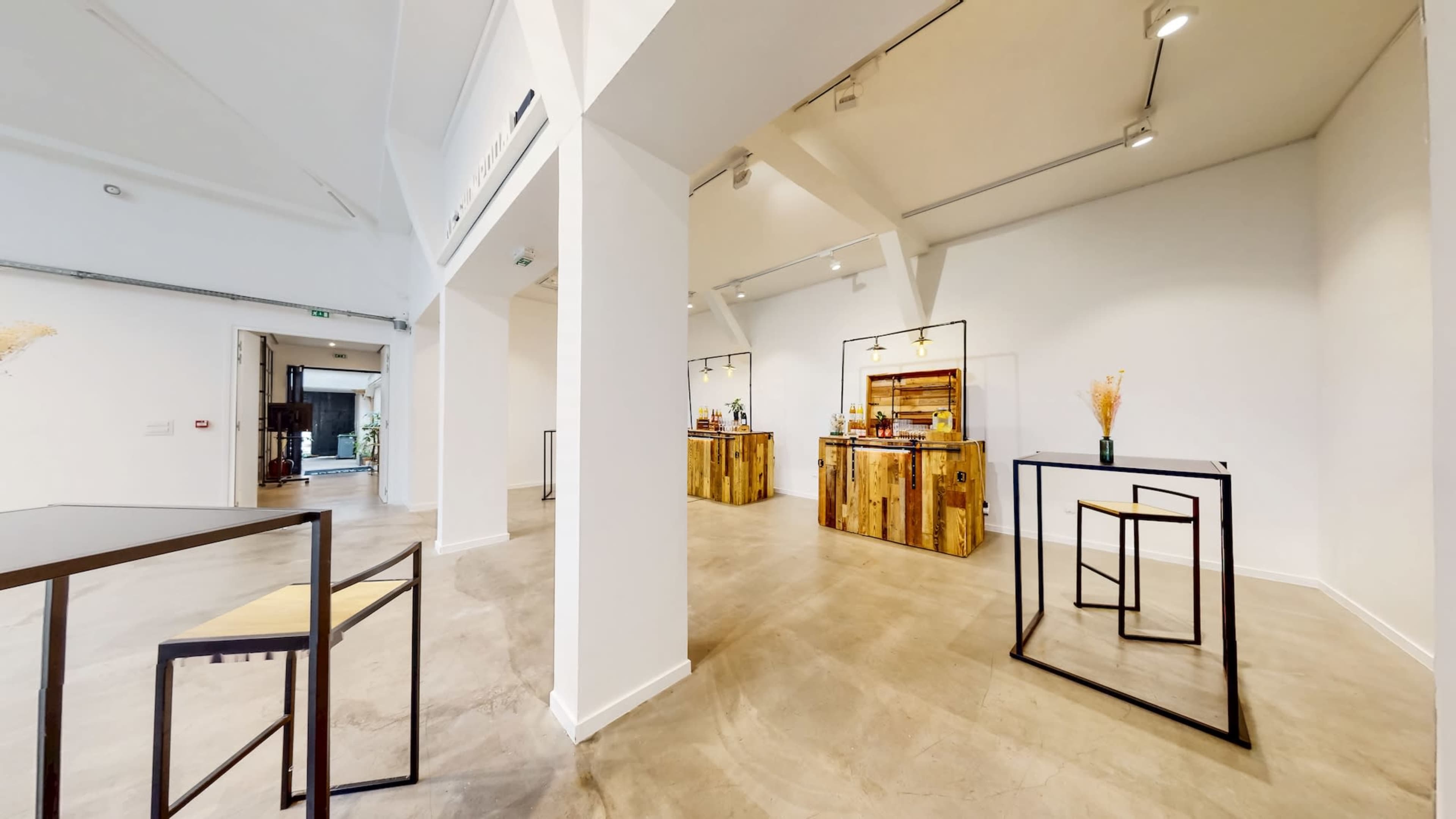 The image shows a spacious, modern interior featuring white walls and concrete flooring, with two wooden service counters in the background and minimalistic furniture arranged throughout.