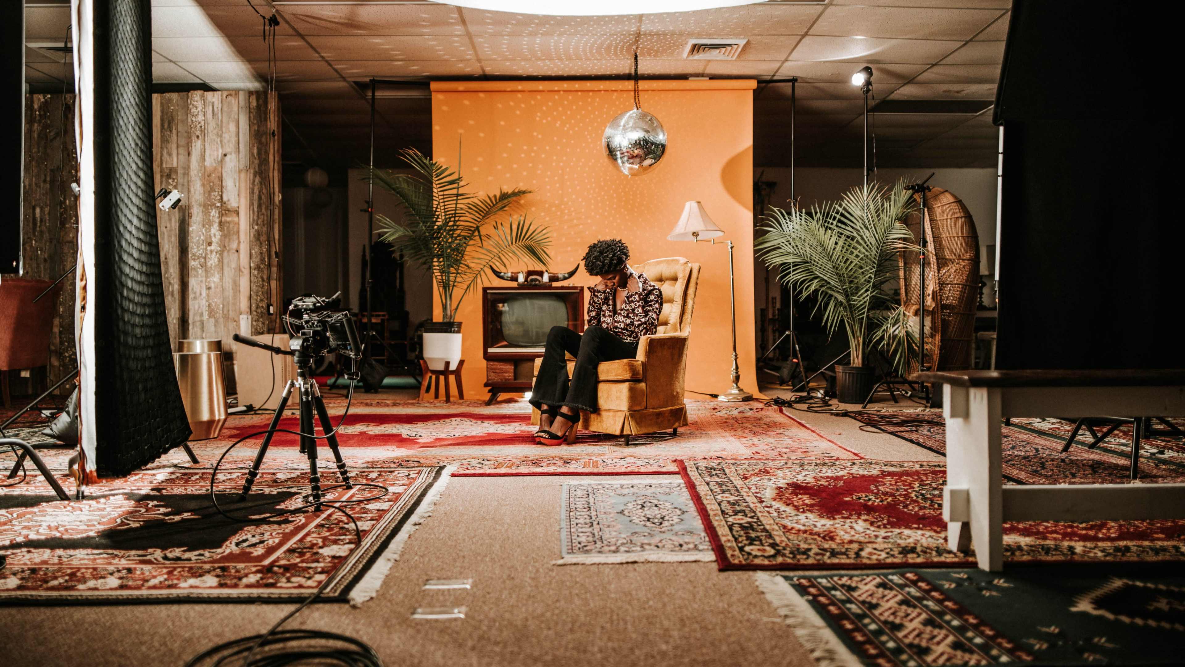 A person sits in a vintage armchair in a studio setting, surrounded by plants, a retro television, and camera equipment on a patterned rug.