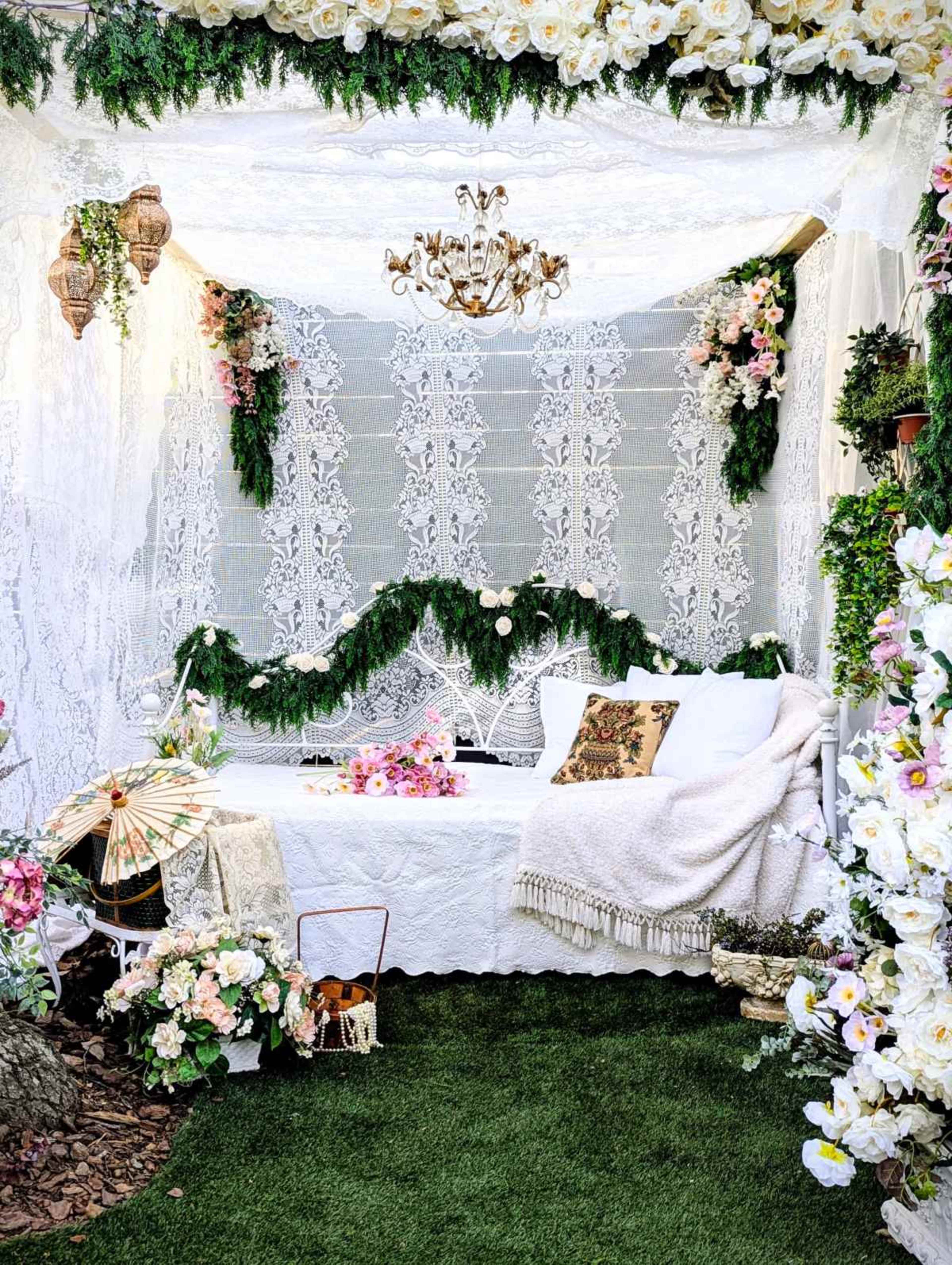 The image shows a cozy, decorated nook surrounded by floral arrangements and lace fabric, featuring a white couch, decorative pillows, and a chandelier overhead.