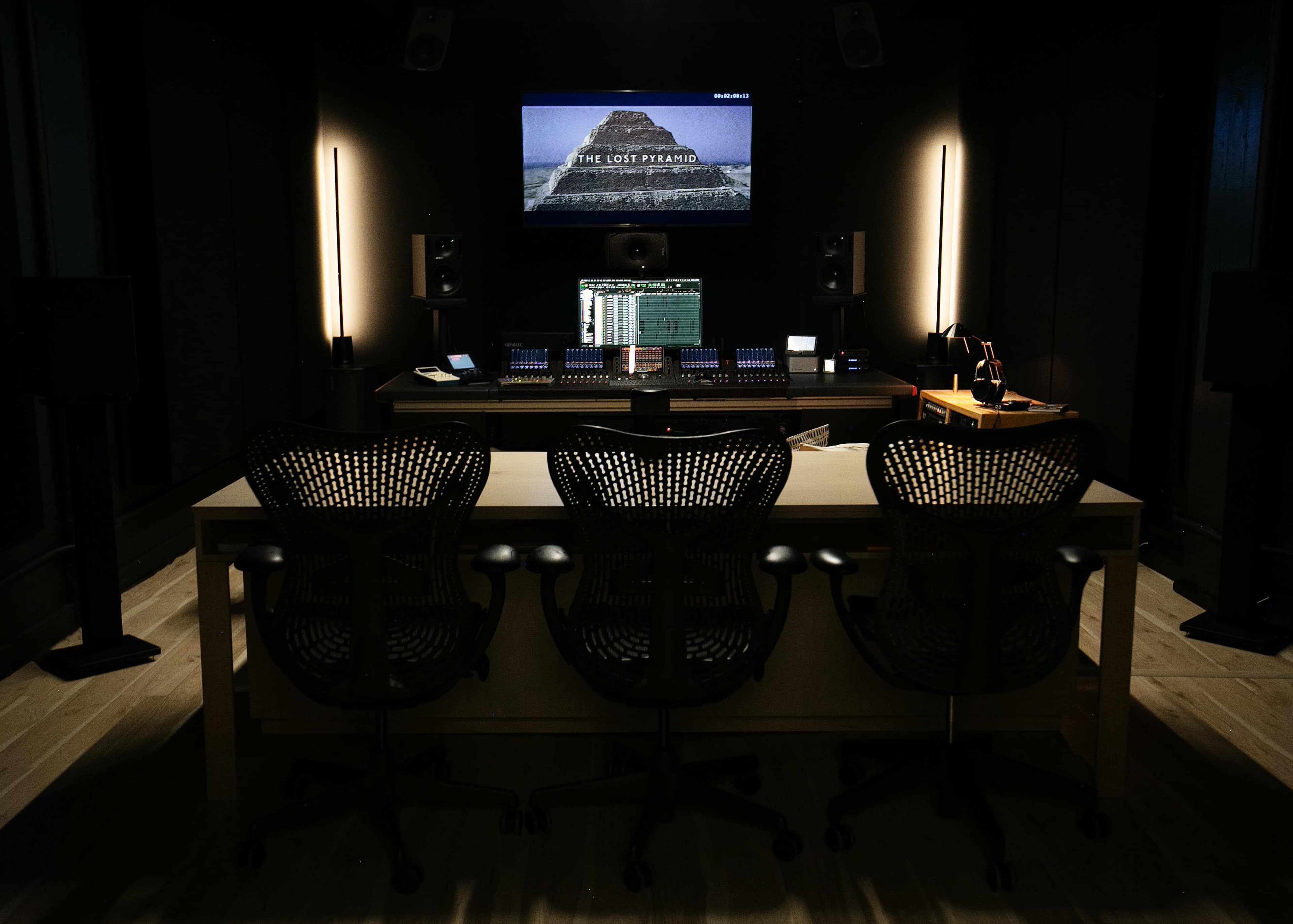 A dark recording studio with a central desk featuring audio equipment, three black office chairs, and a screen displaying "THE LOST PYRAMID."