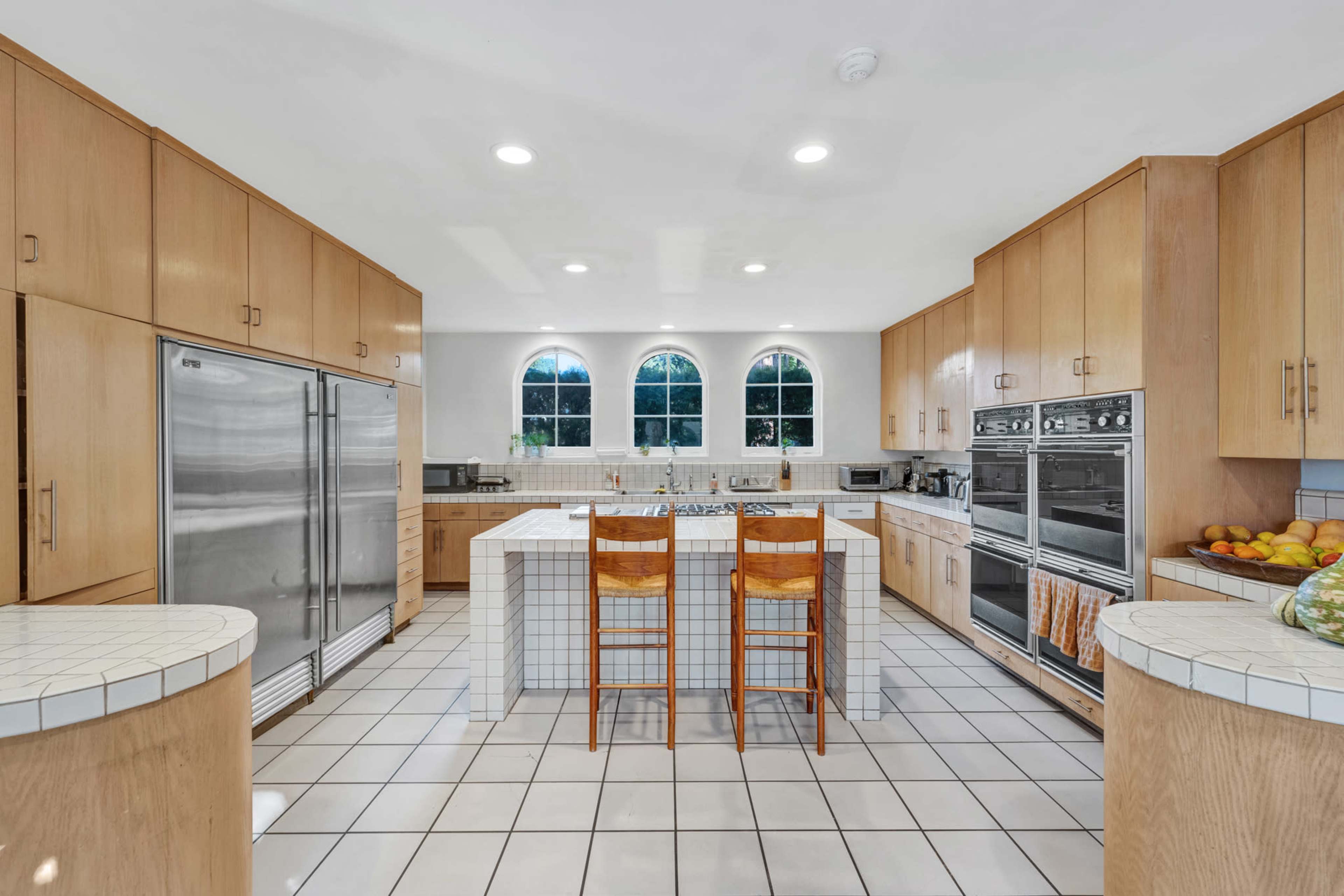 A spacious kitchen features light wood cabinetry, stainless steel appliances, and a tiled floor with two wooden stools at the center island.