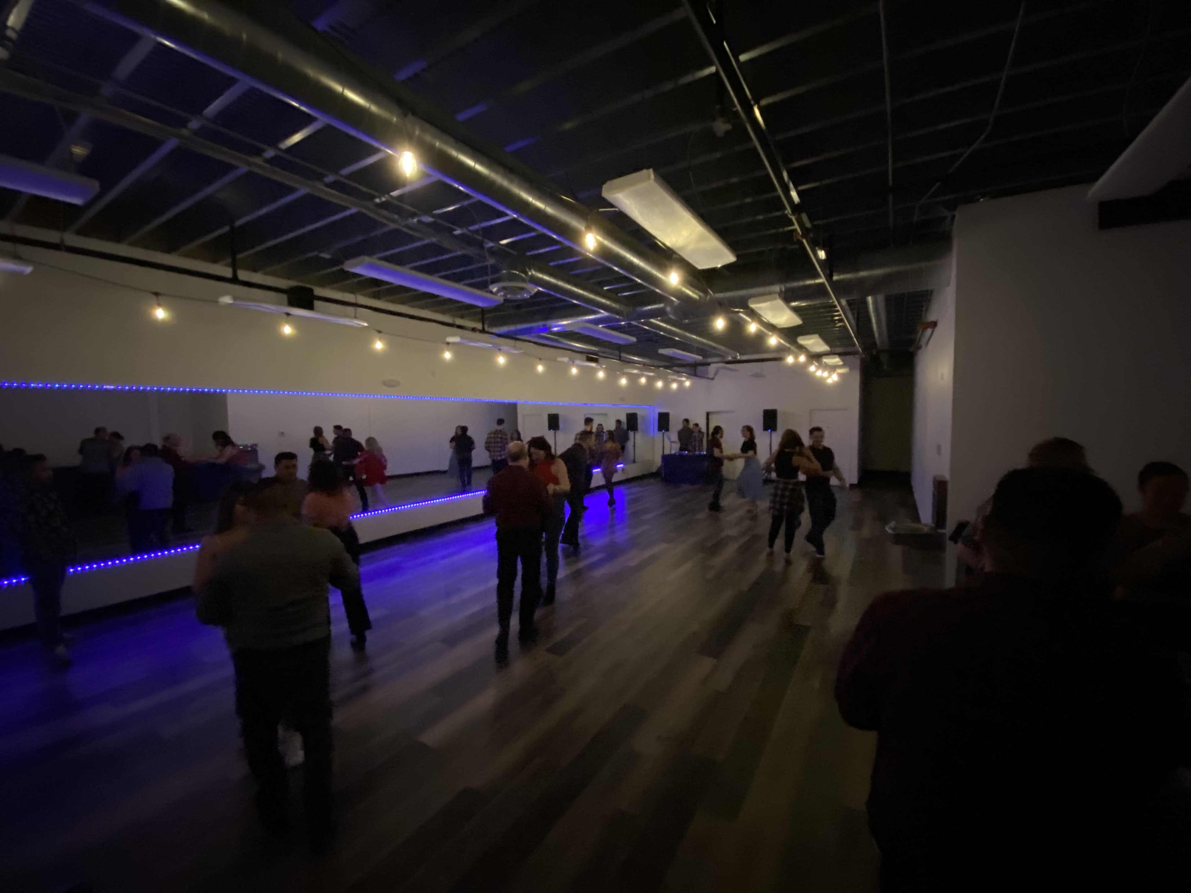 A dance studio with wooden flooring and blue lights, featuring several people dancing near a mirrored wall.