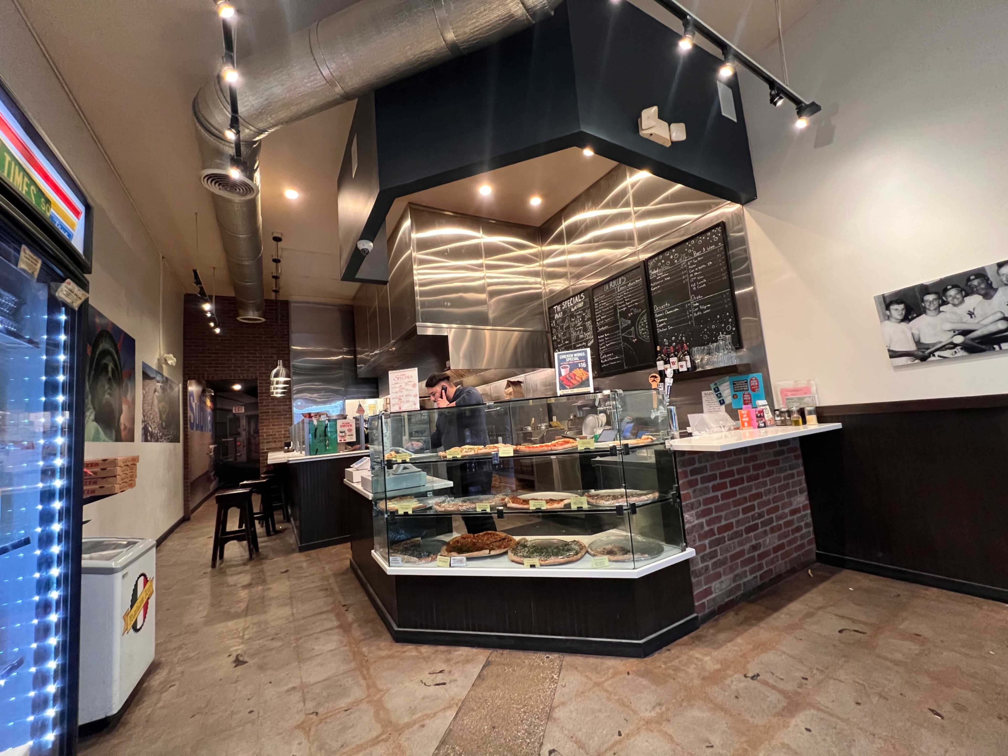 The image shows a modern dessert shop featuring a display case filled with various pastries and a barista preparing orders behind the counter.