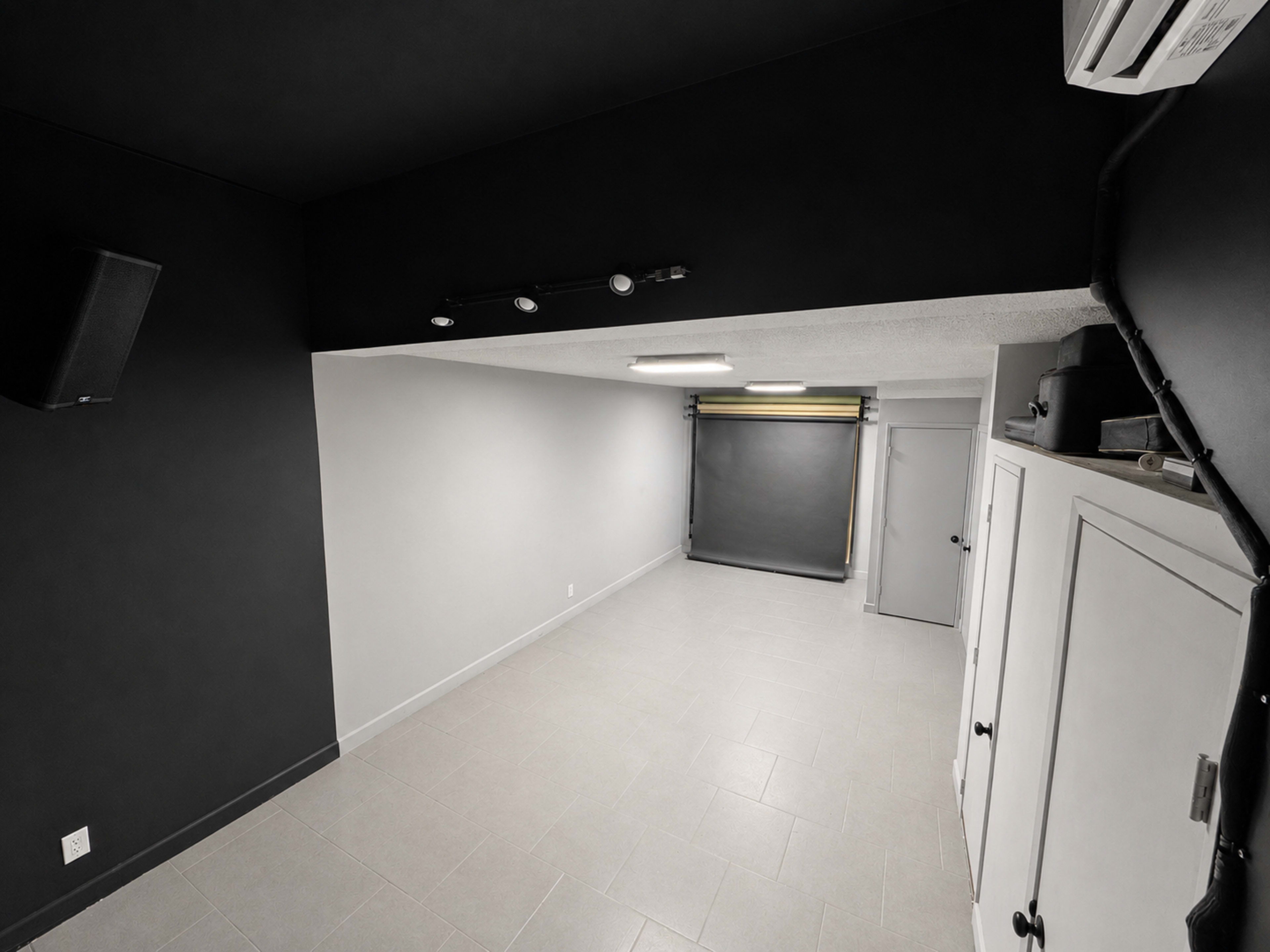 The image shows a minimally furnished room with dark walls, light tile flooring, and a closed roll-up shade at one end.