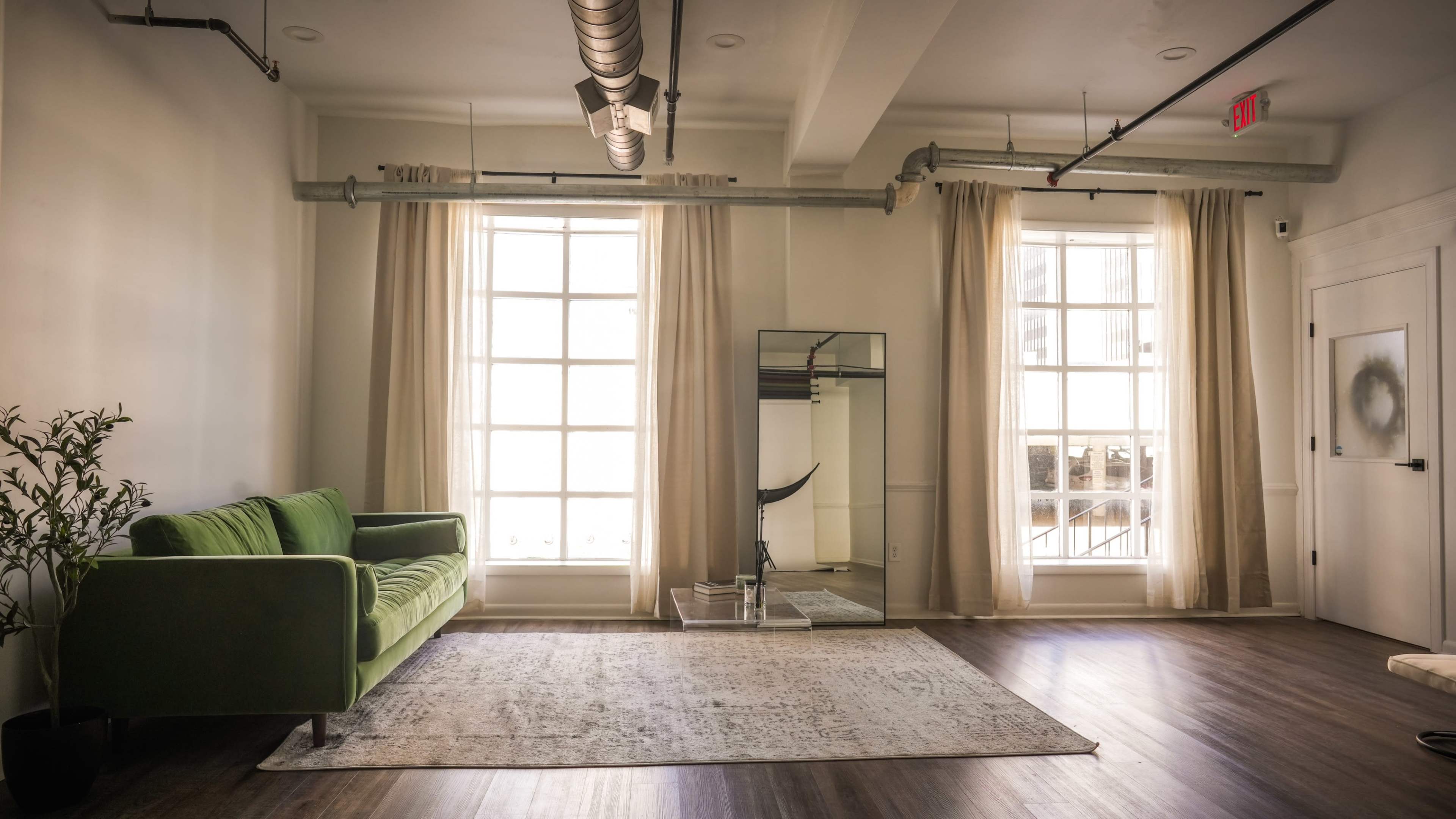 A bright room features a green sofa, large windows with sheer curtains, a mirror, and a patterned rug on a wooden floor.