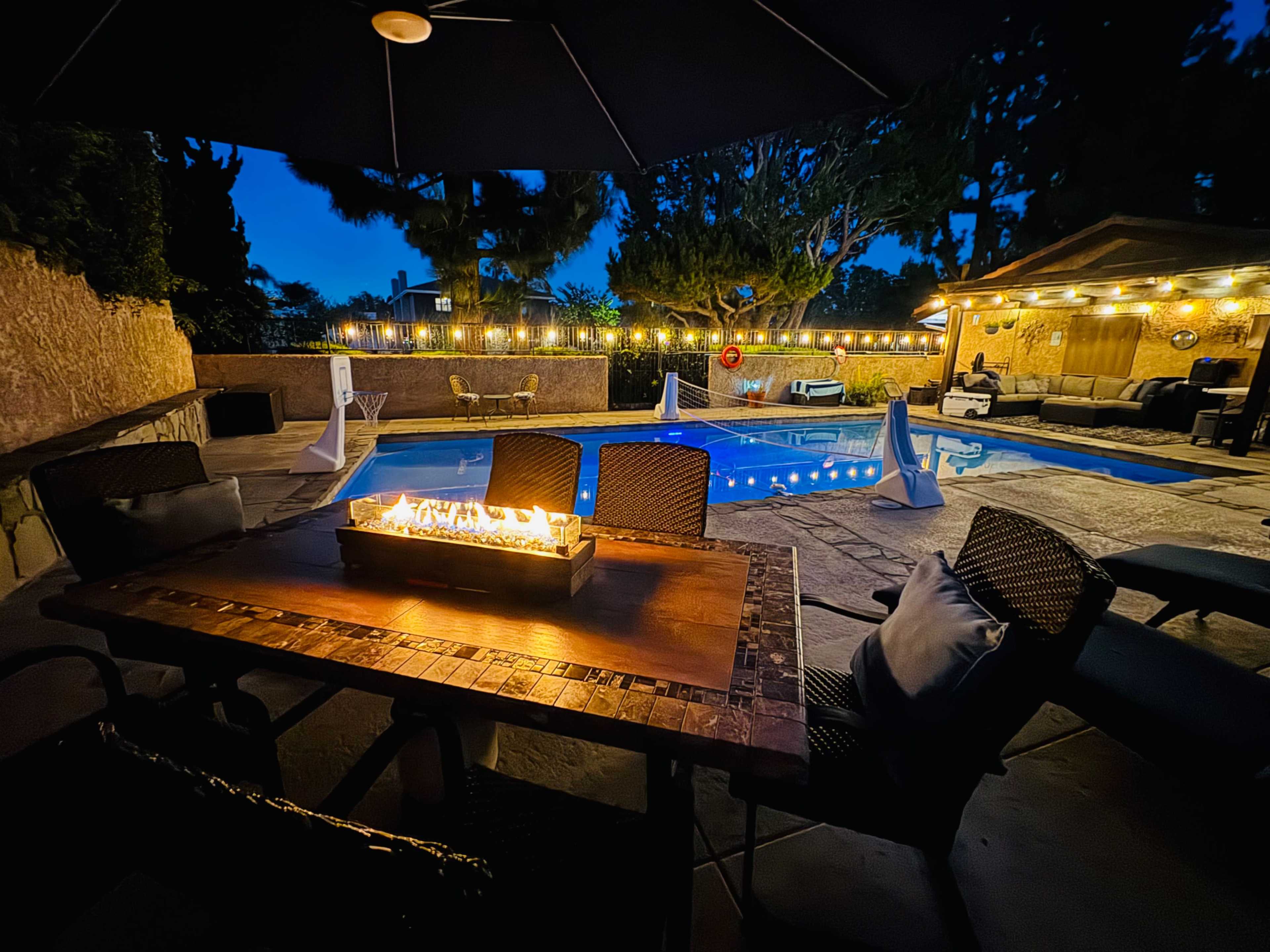 An outdoor dining area with a fire feature overlooks a lit swimming pool surrounded by trees at dusk.