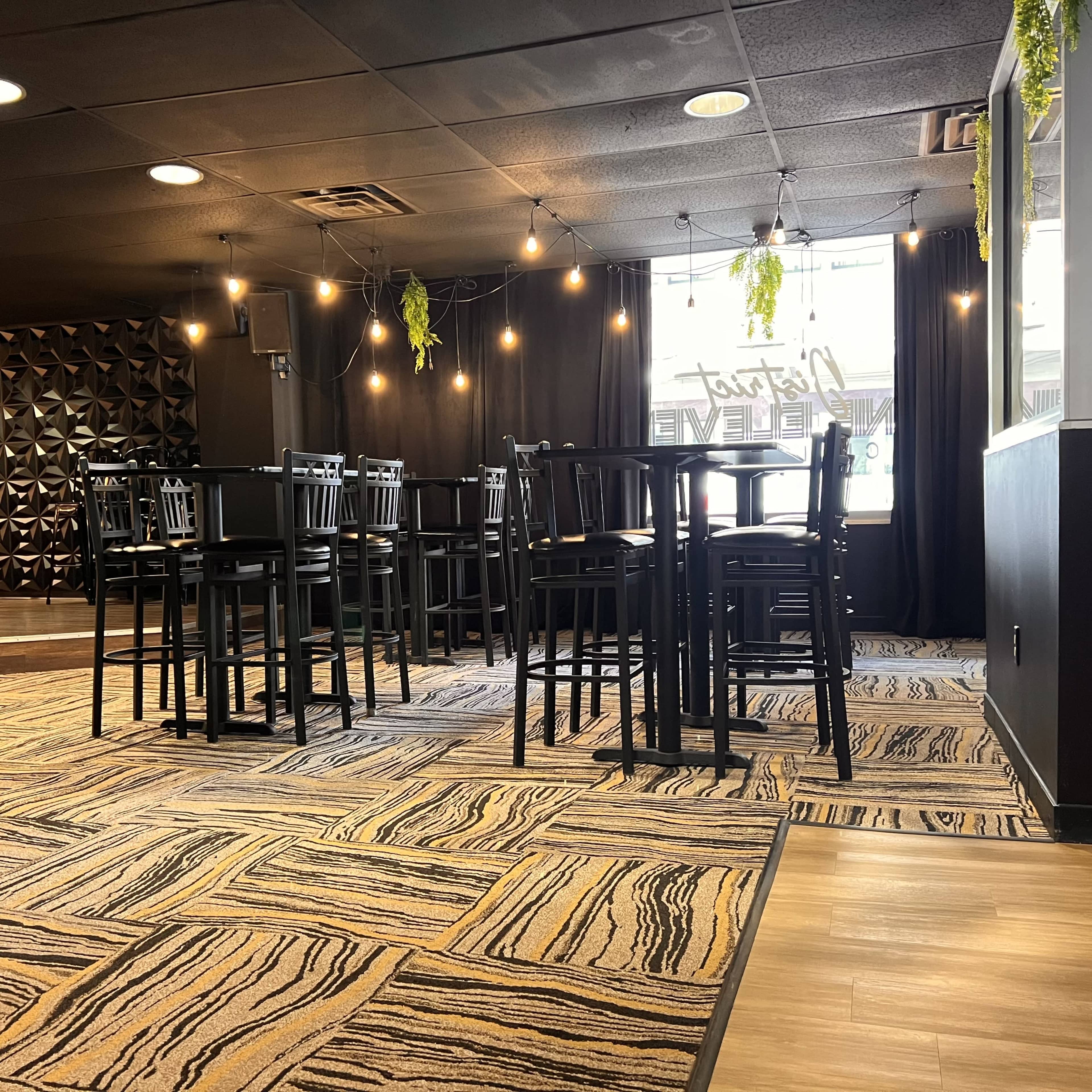 The image shows a dimly lit dining area with black wooden tables and chairs, a patterned carpet, and decorative hanging plants.