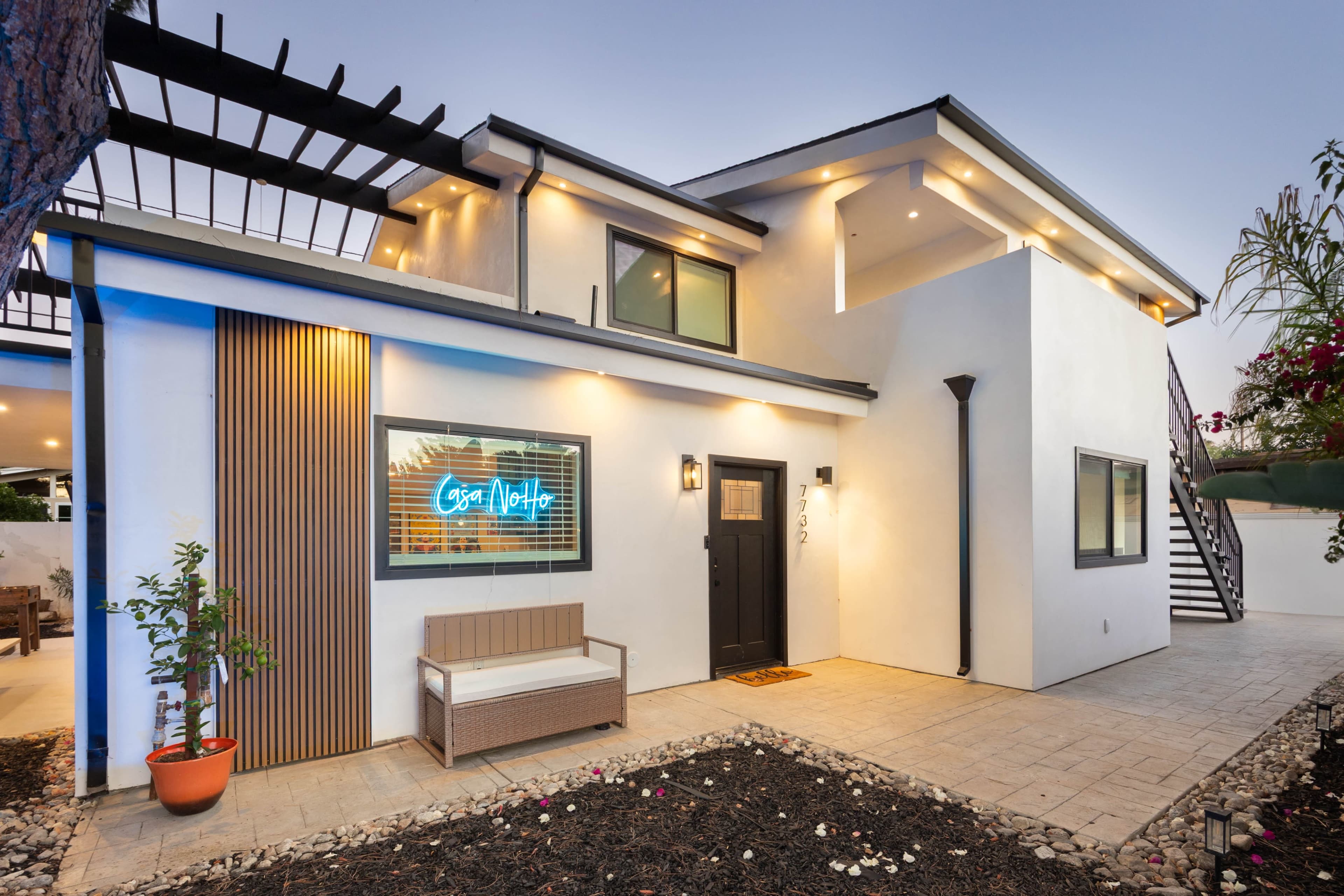 The image shows a modern, two-story house with a front porch featuring a bench and a brightly lit neon sign.