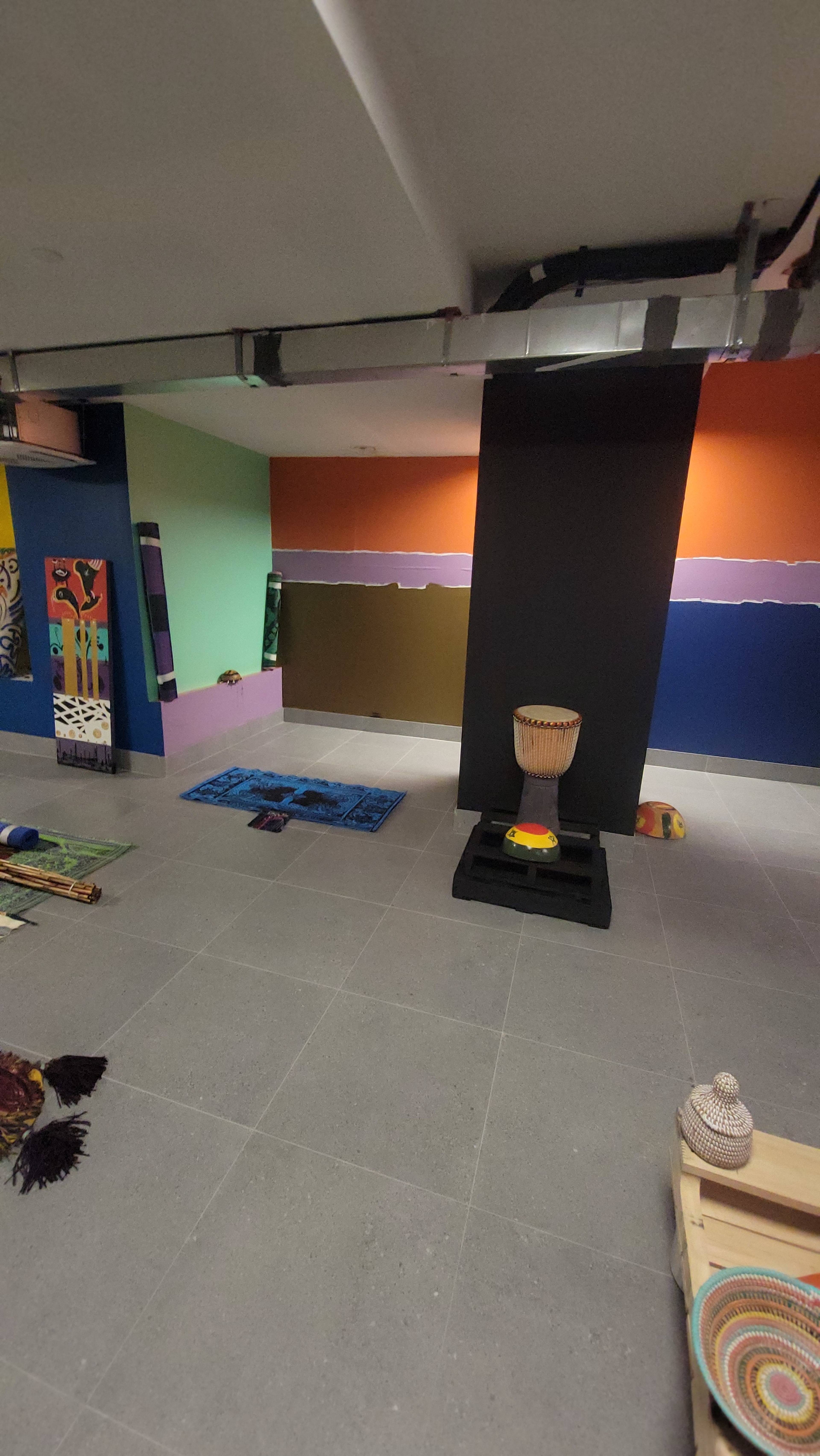 The image shows a colorful indoor space with various textured rugs on the floor, a large black column, and a decorative drum placed on a black stand.