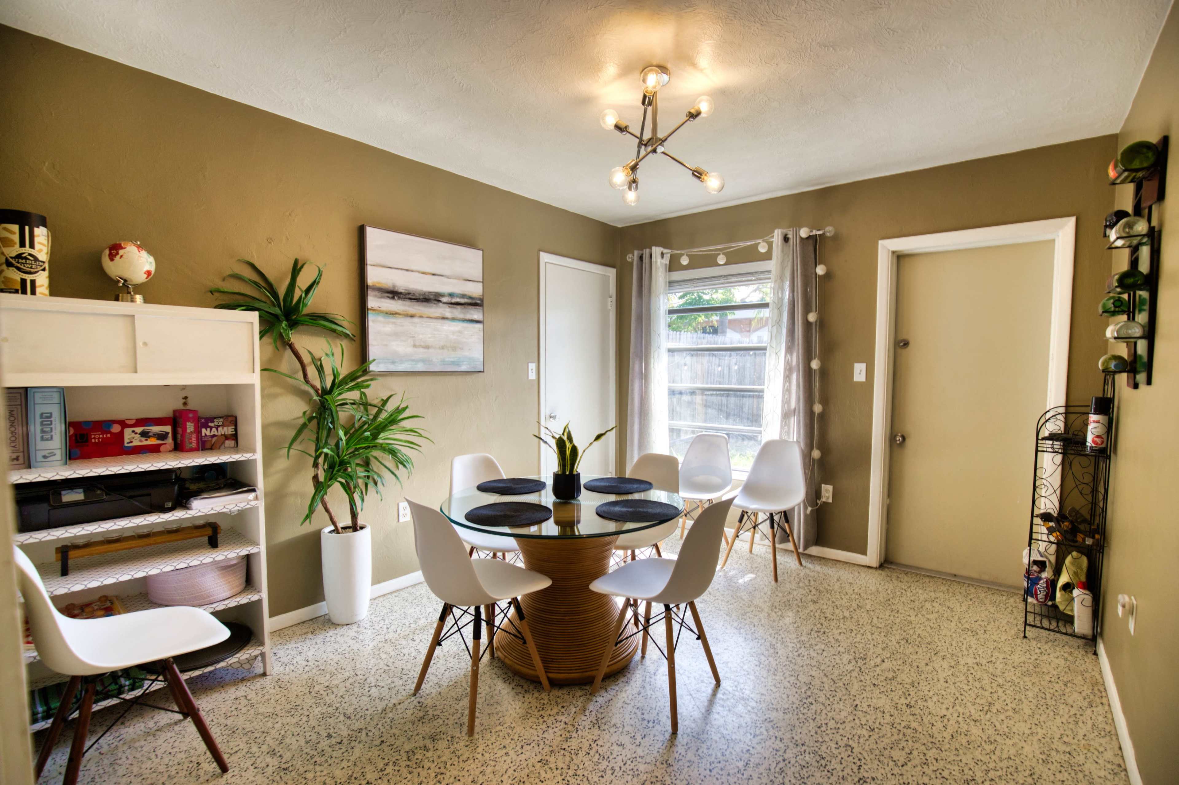 A modern dining area features a round table surrounded by white chairs, a potted plant, and a small shelf with decorative items.