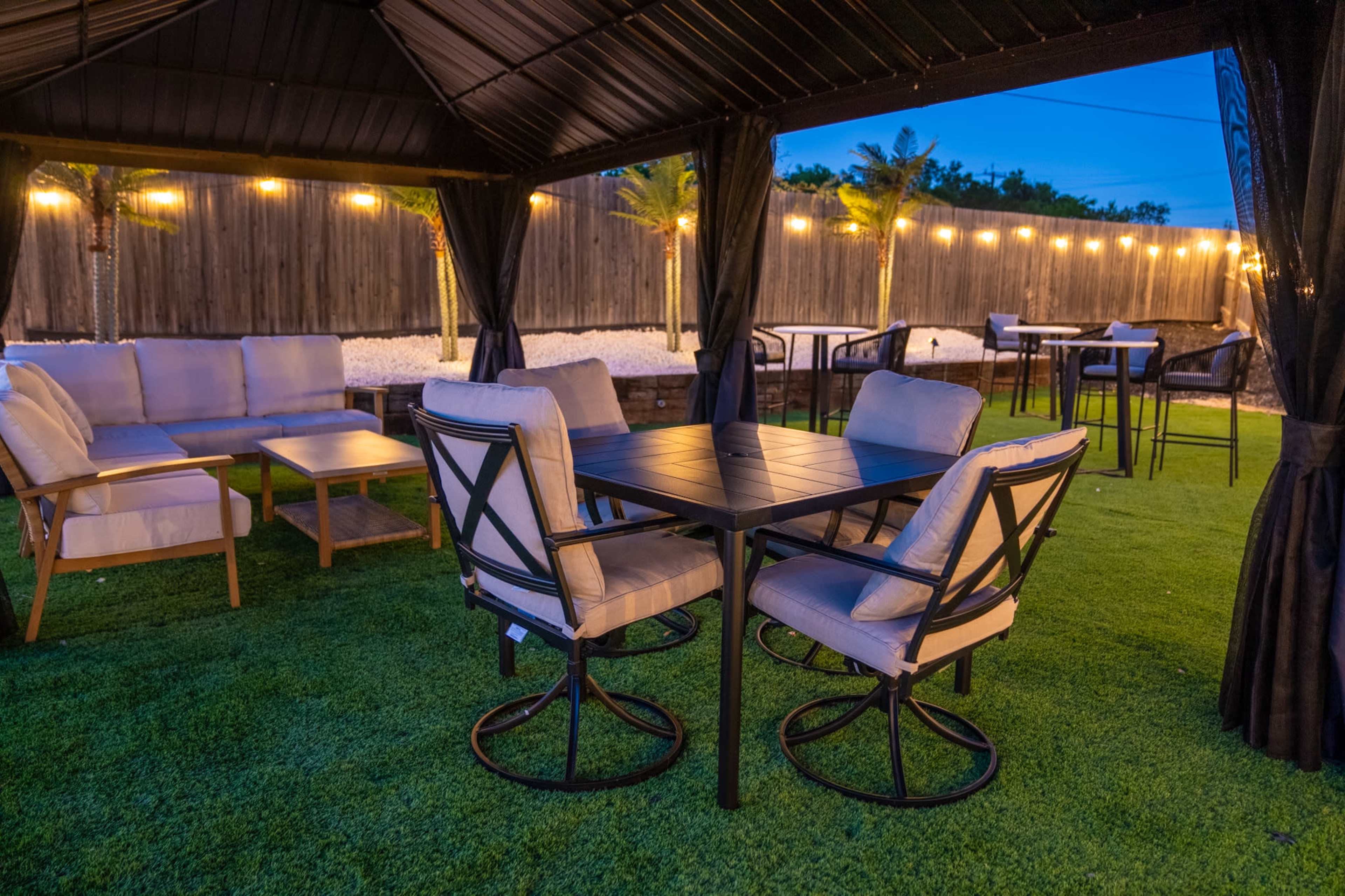 A canopied outdoor seating area features a table surrounded by swivel chairs, with sofas placed to one side and string lights illuminating the space.