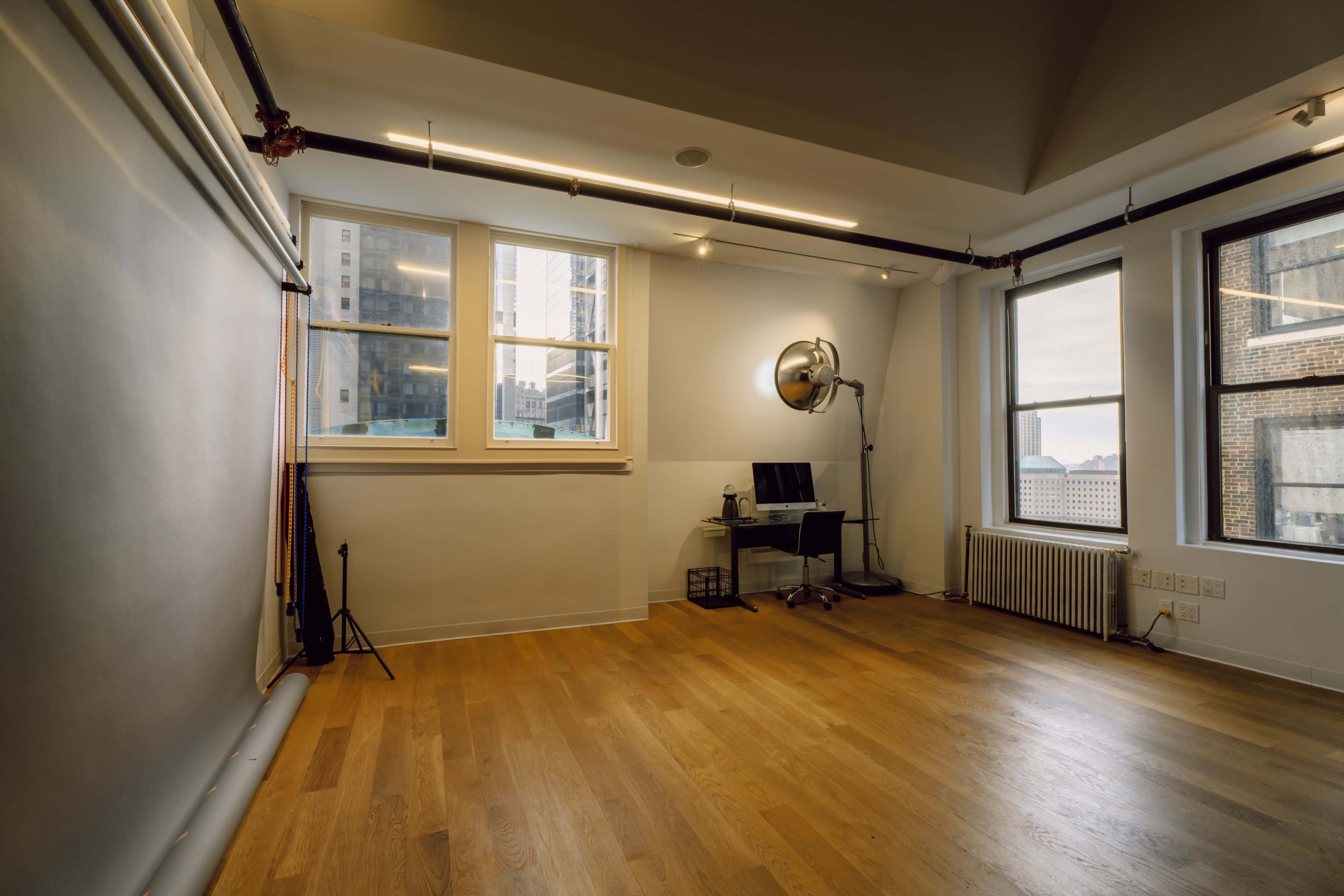 A spacious, well-lit room with large windows, wooden flooring, and a simple setup including a backdrop, a computer desk, and lighting equipment.