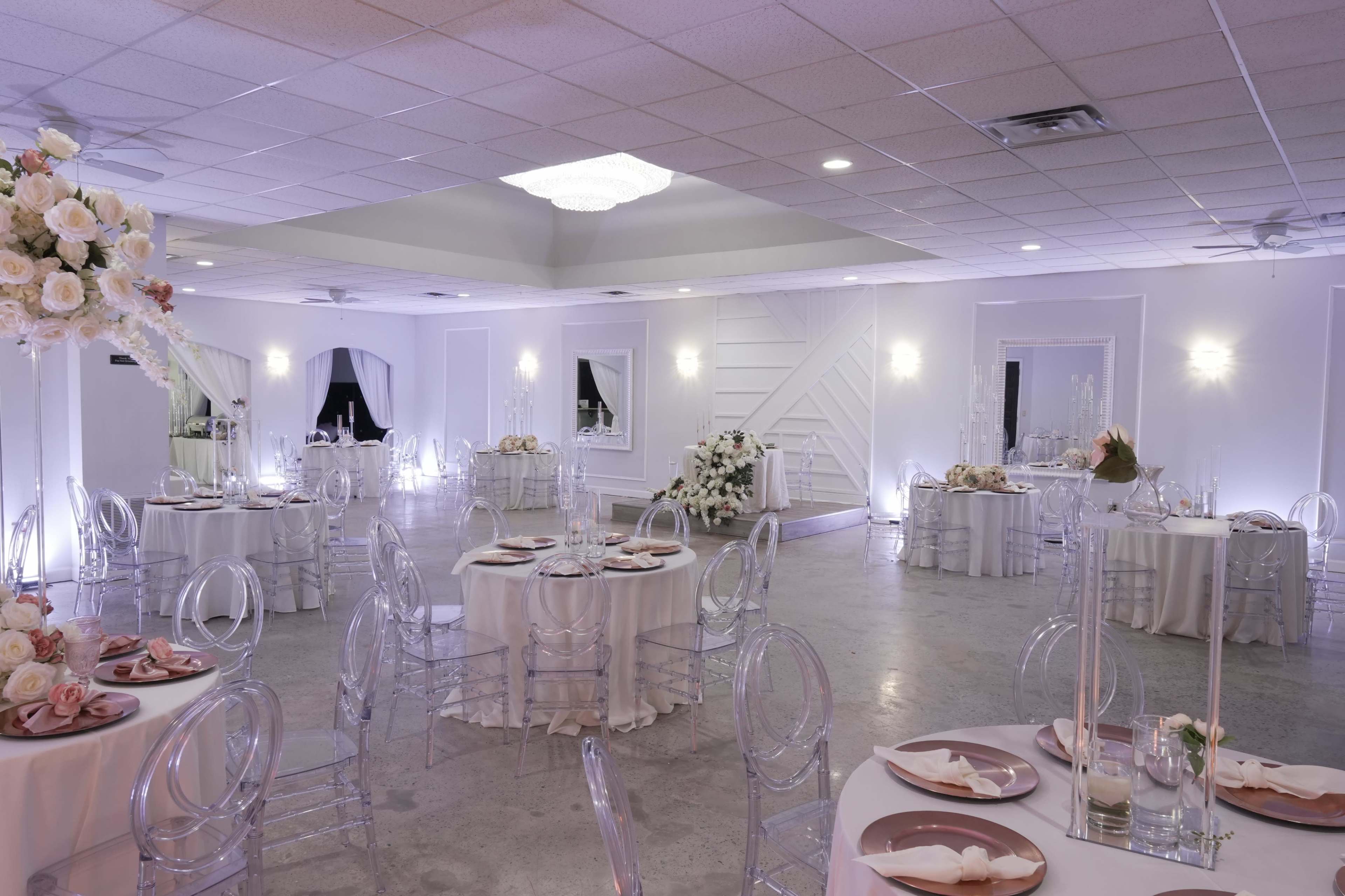 A well-decorated banquet hall features round tables with elegant white linens, clear chairs, and floral centerpieces, illuminated by soft lighting.