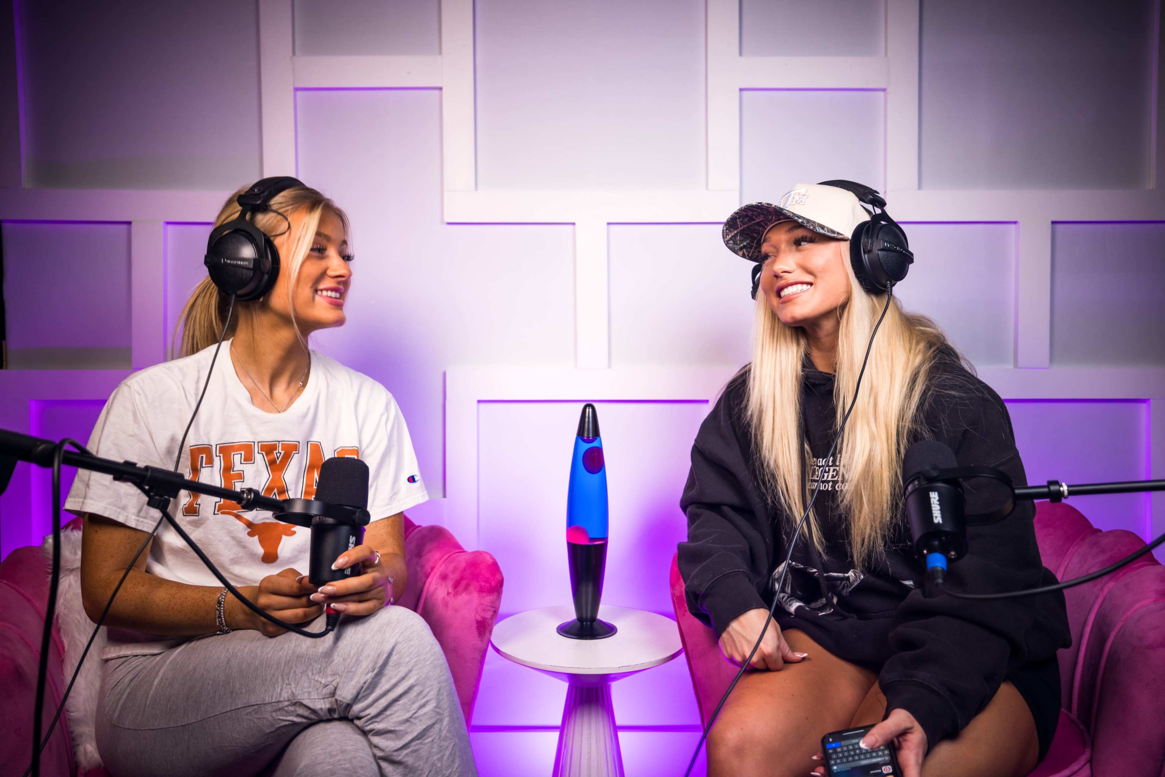 Two women wearing headphones sit on pink chairs in a studio with a purple-lit backdrop, engaging in conversation while microphones are positioned in front of them.