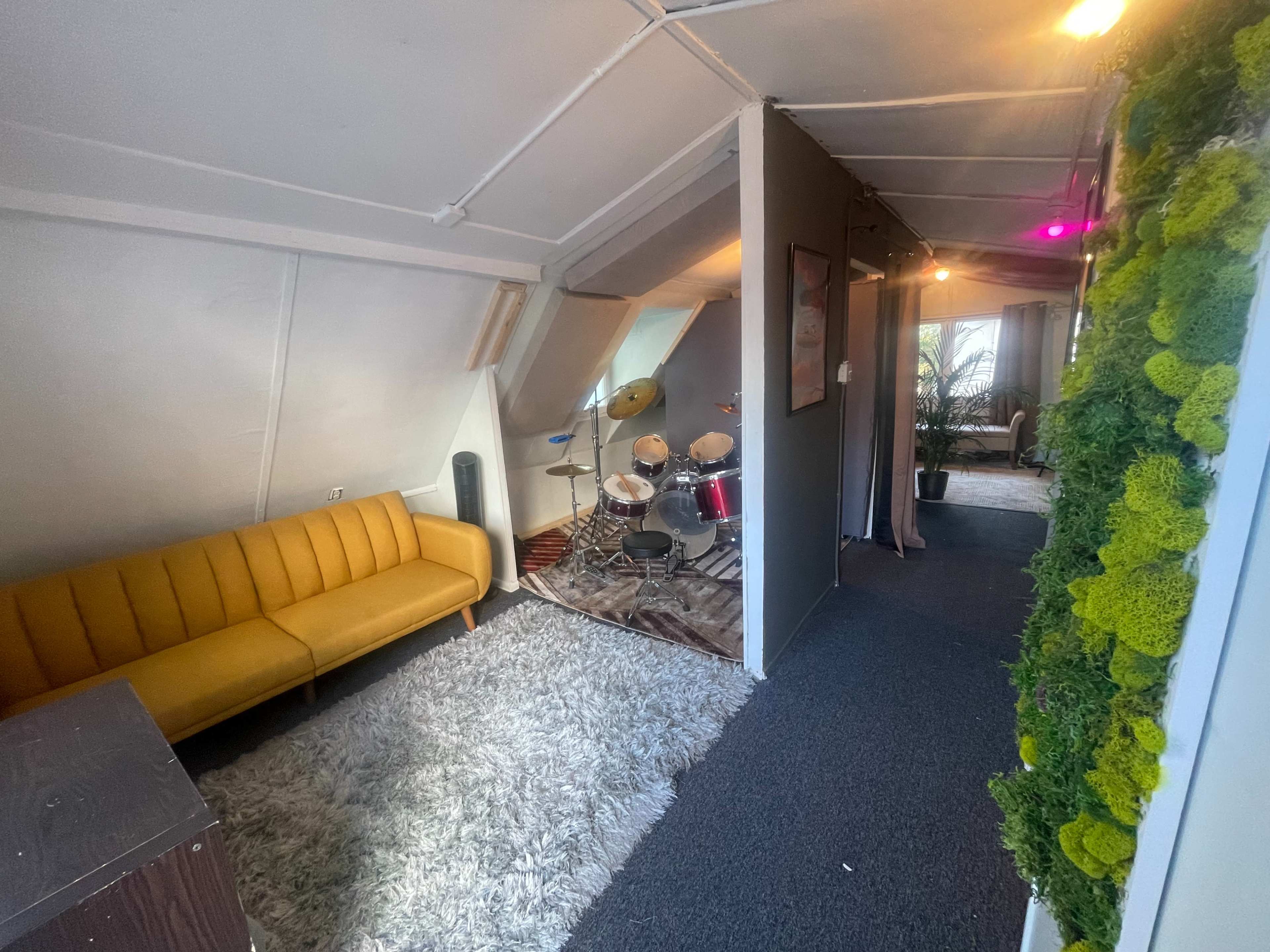 A small room with a yellow couch on a carpeted floor, leading to a space with a drum set and a plant in the corner.