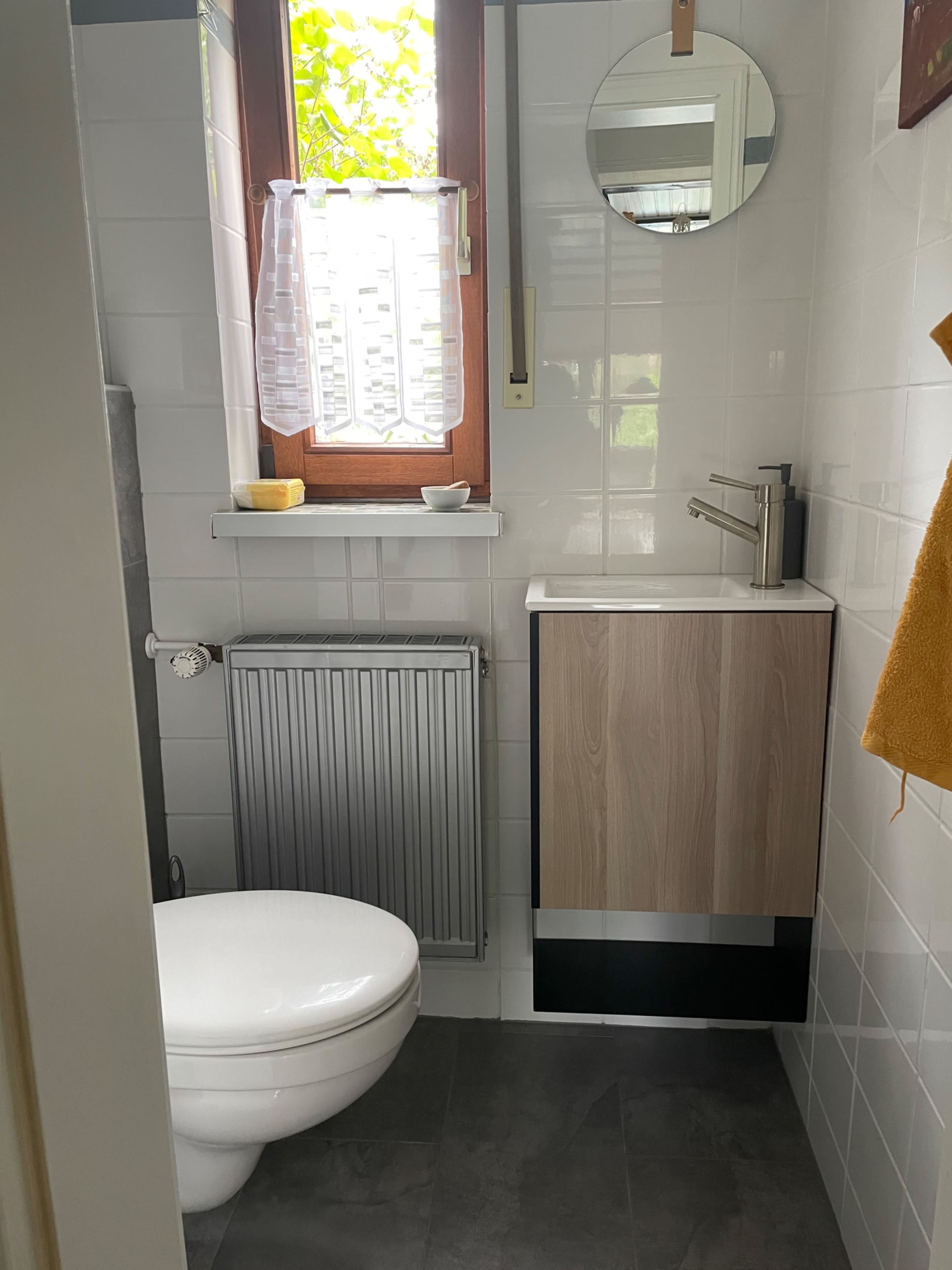 A small bathroom featuring a white toilet, a wall-mounted sink with a wooden cabinet, a mirror, and a window with light filtering through.