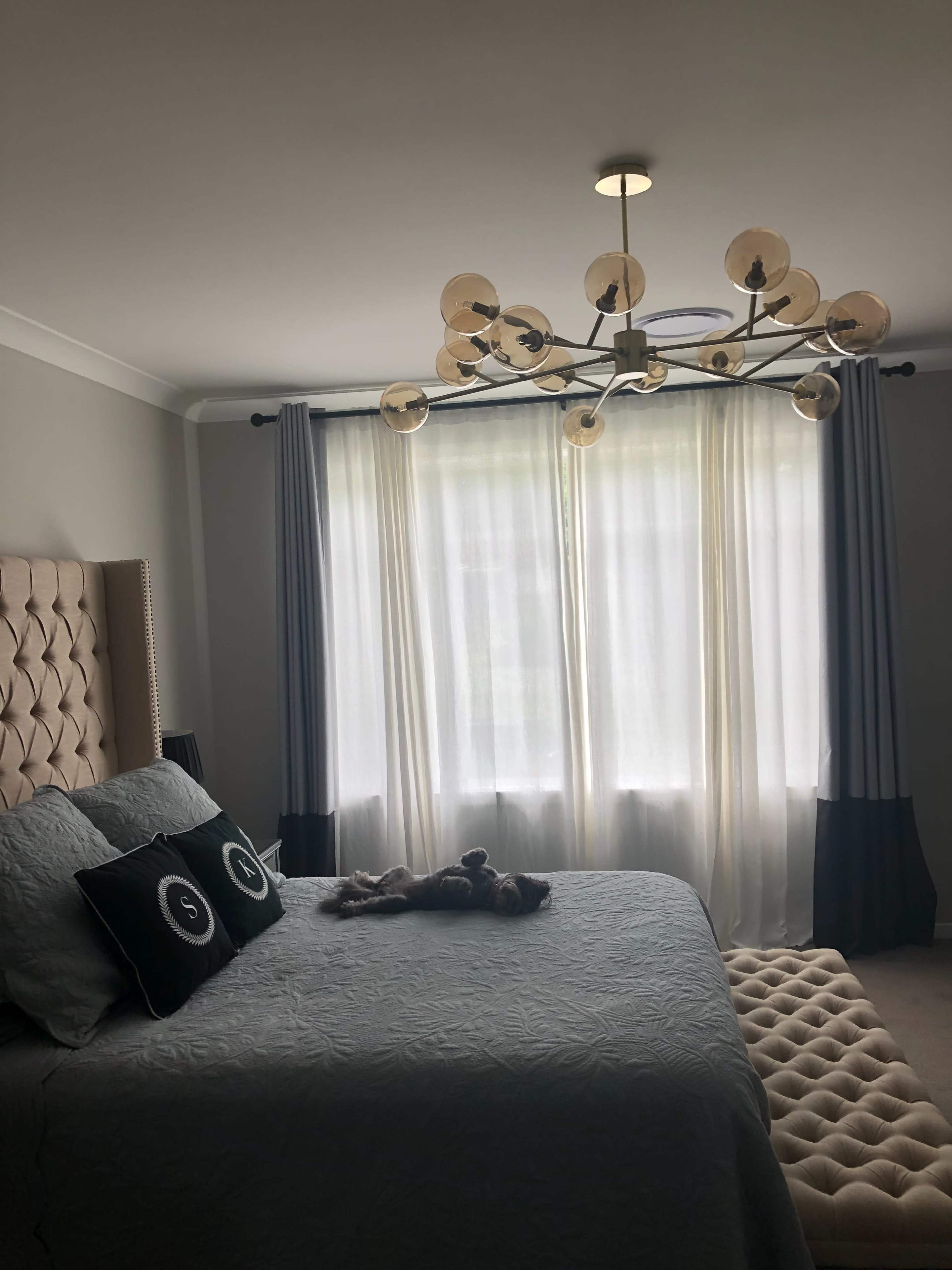 The image shows a well-decorated bedroom featuring a large bed with a tufted headboard, a chandelier, and long curtains framing a window.