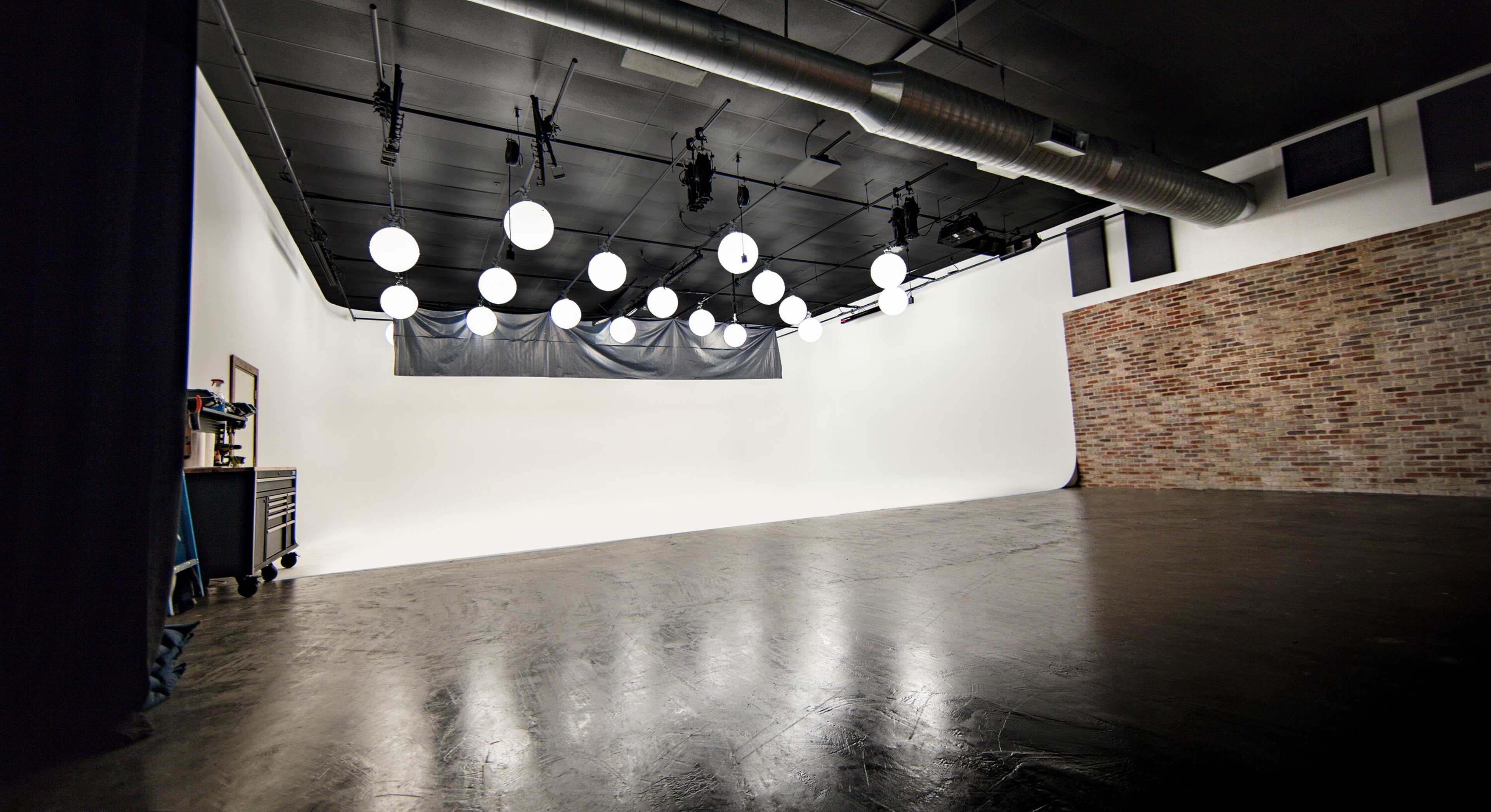A photography studio with a blank white backdrop, dimly lit by multiple round lights hanging from the ceiling, and a brick wall on one side.