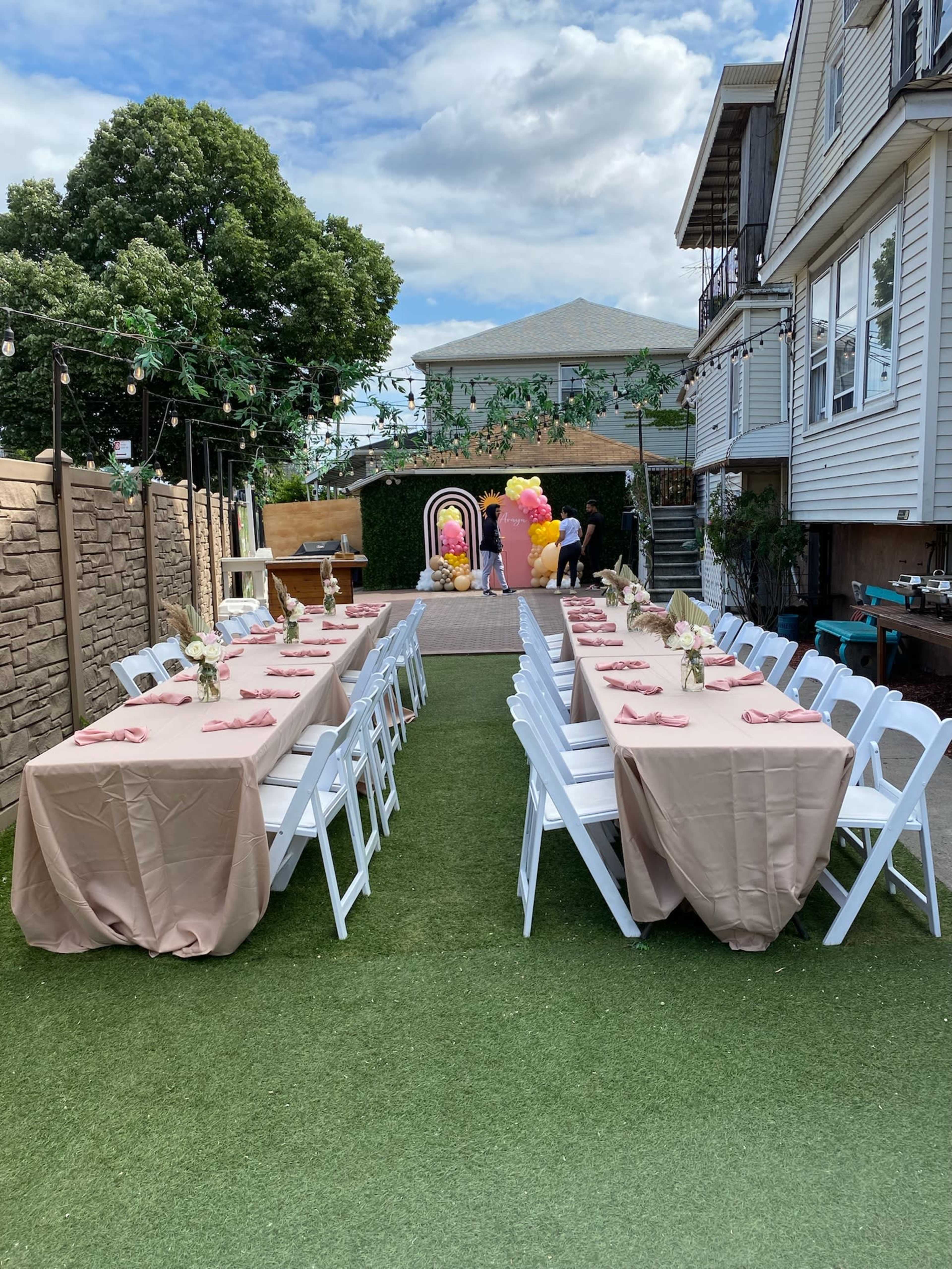 A neatly arranged outdoor event space with long tables covered in beige tablecloths, set with pink napkins, surrounded by greenery and decorations.