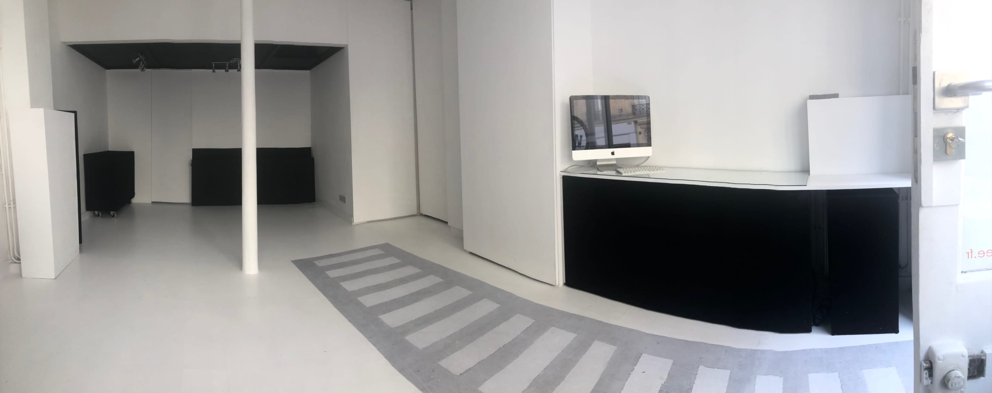A minimalist, white-walled interior space featuring a reception area with a computer, a gray striped pedestrian walkway on the floor, and two black furniture pieces in the background.