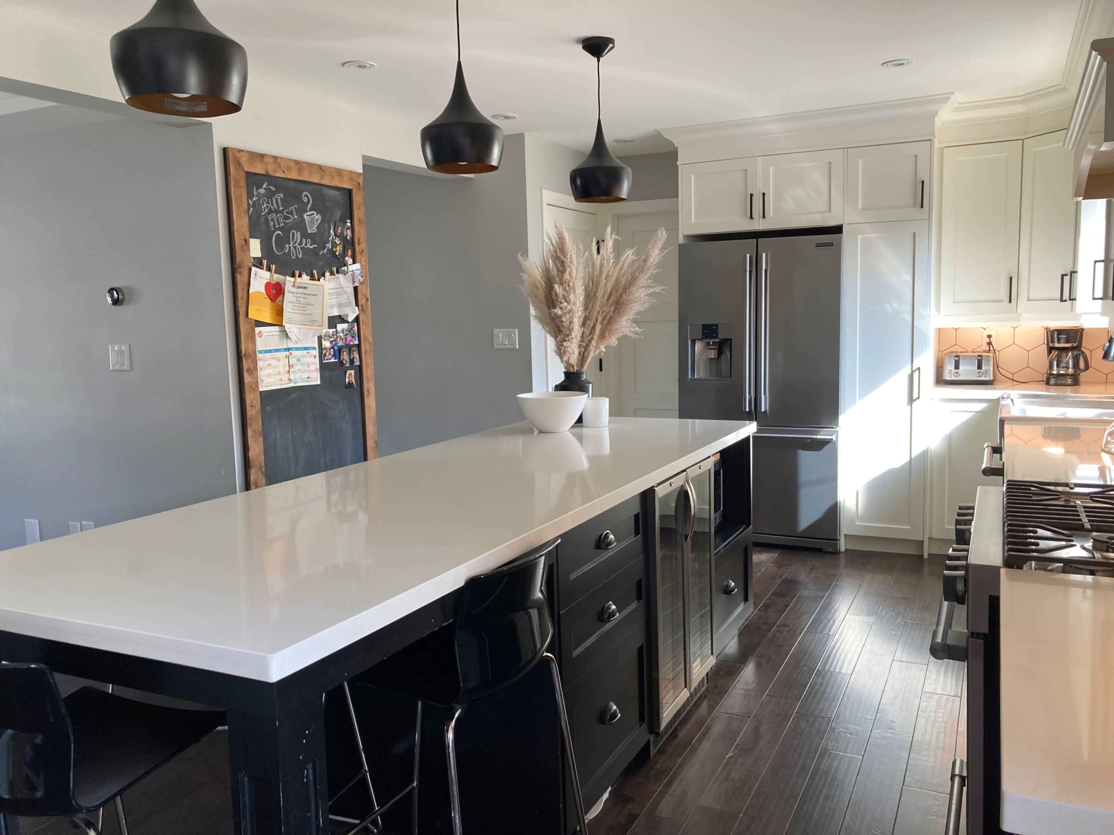 A modern kitchen features a large white island with black chairs, stainless steel appliances, and a vase of decorative grasses.