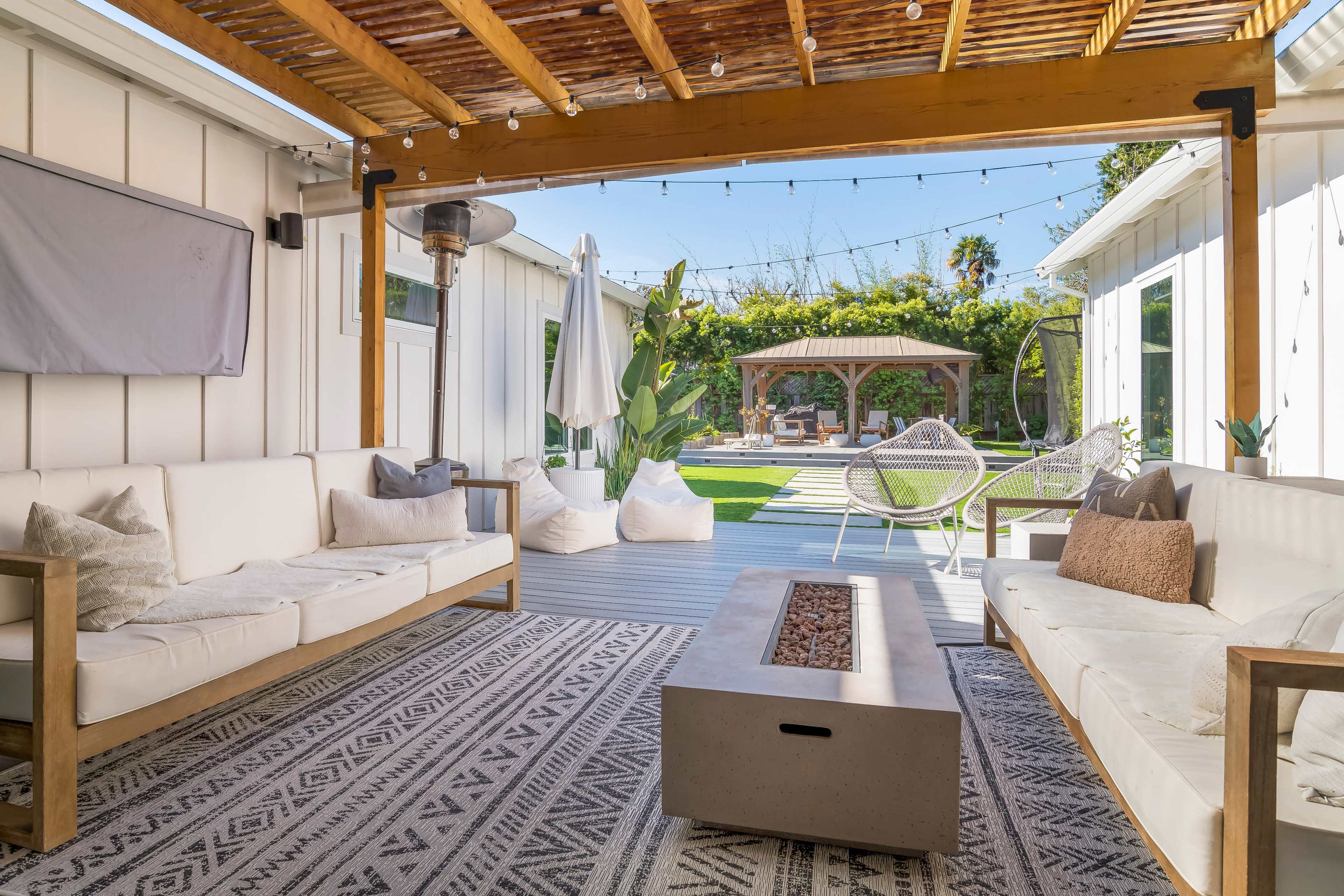 The image shows a stylish outdoor living space with comfortable white sofas, a central fire pit, decorative plants, and a pergola overhead.