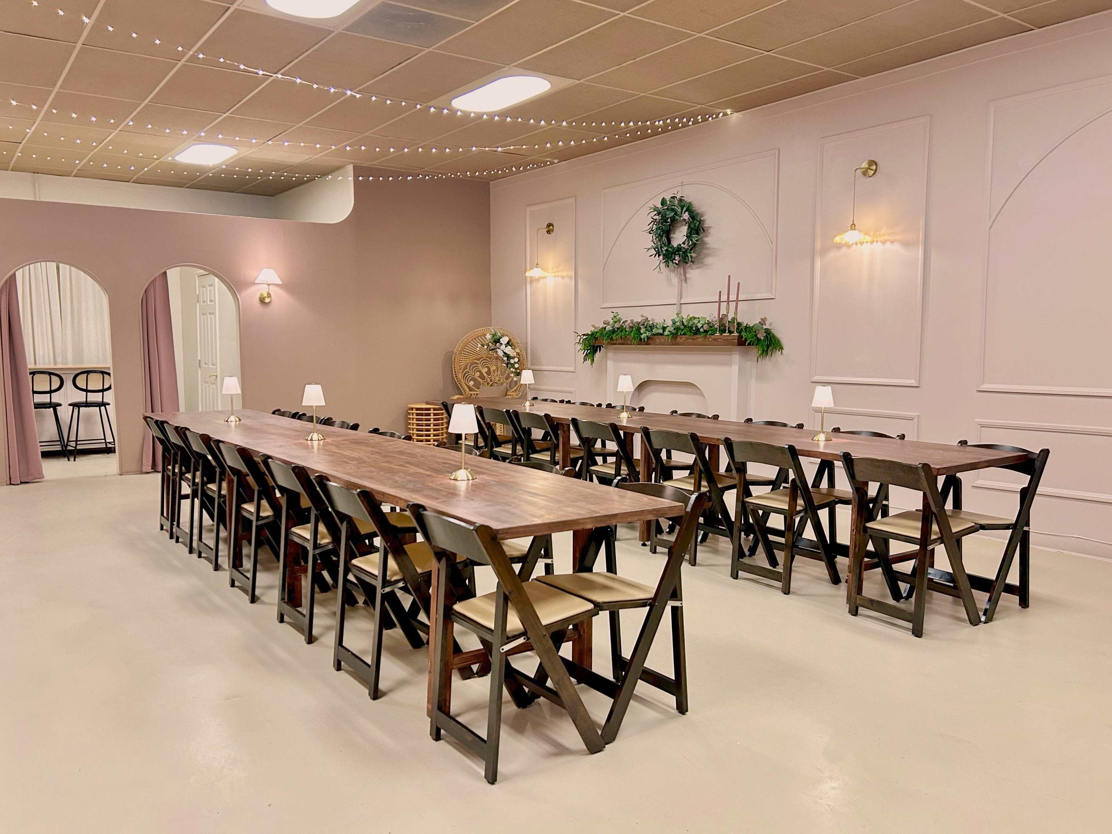 The image shows a large rectangular dining table set with chairs in a softly lit room decorated with wreaths and string lights.