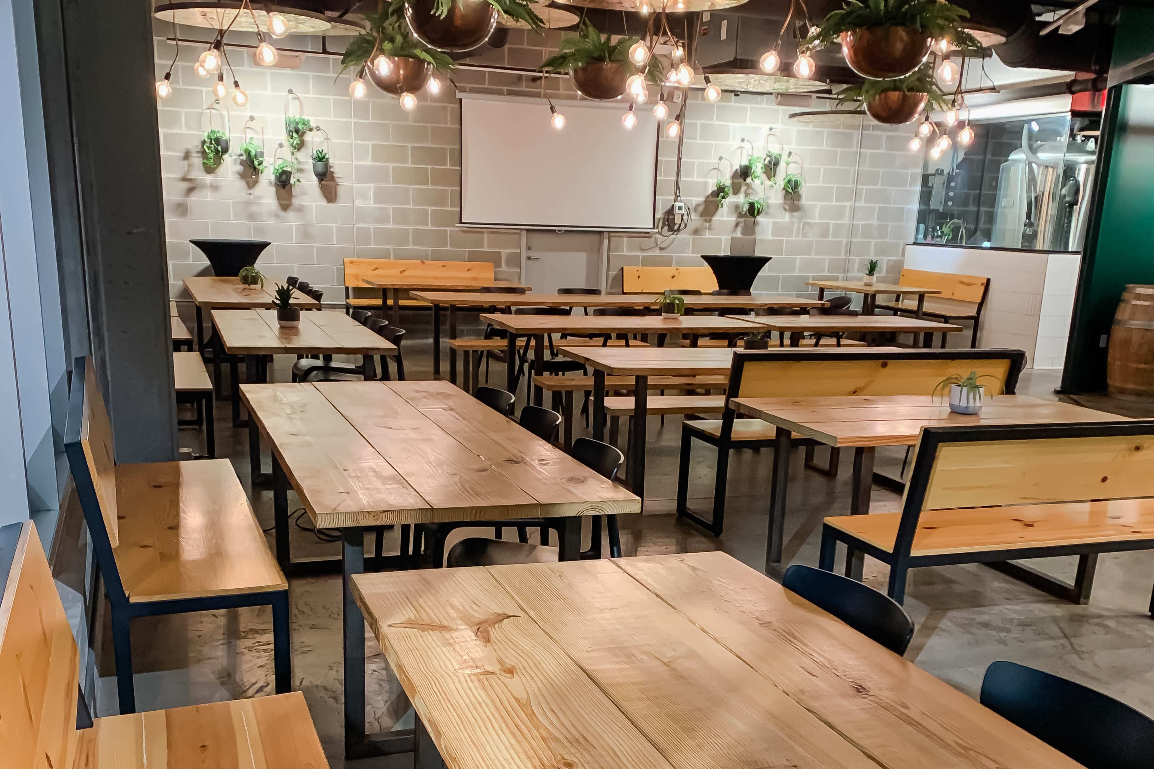 The image shows a spacious dining area with wooden tables and benches arranged neatly, accented by hanging lights and plants.