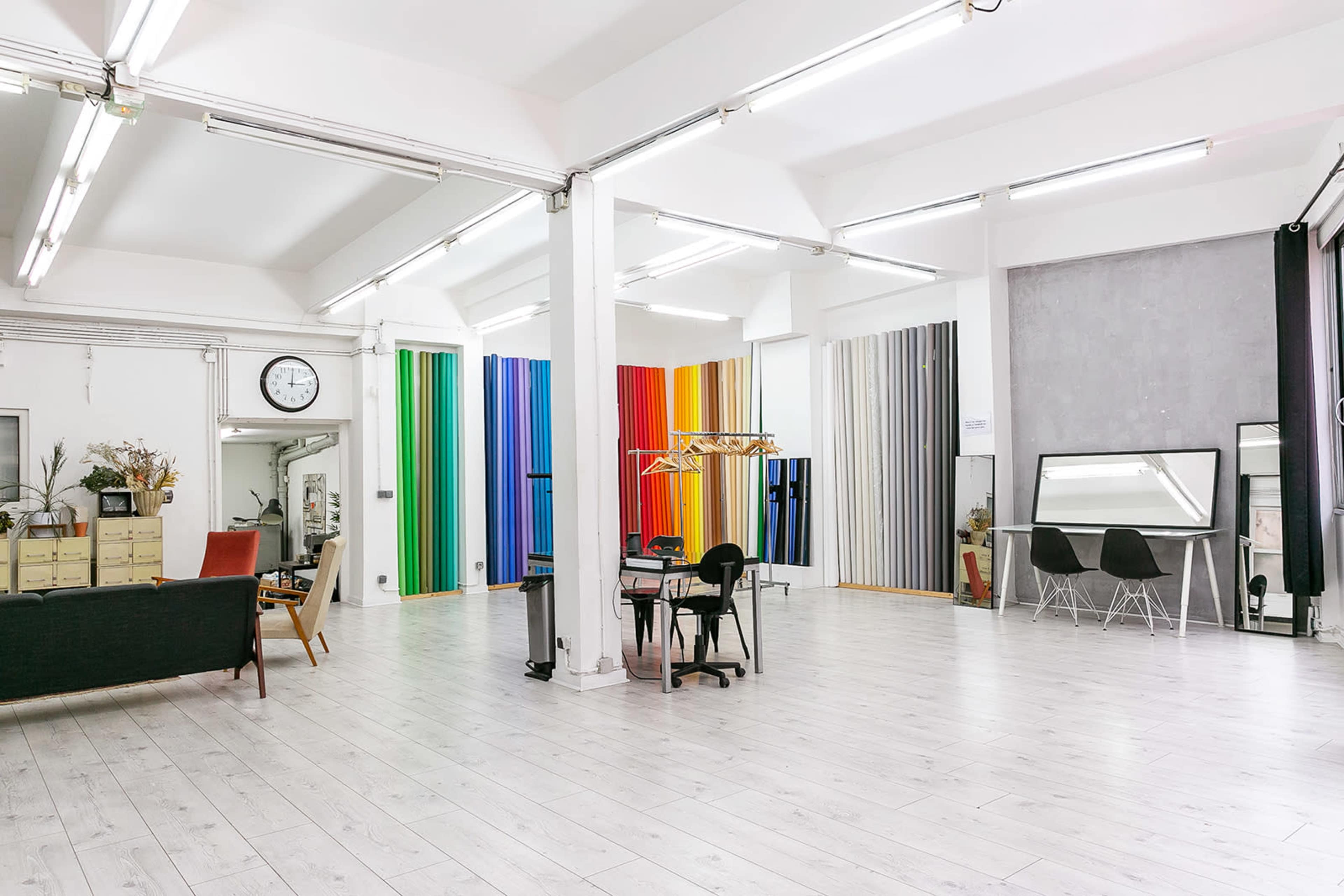 The image shows a brightly lit studio space with colorful vertical fabric panels and a seating area with a clock on the wall.